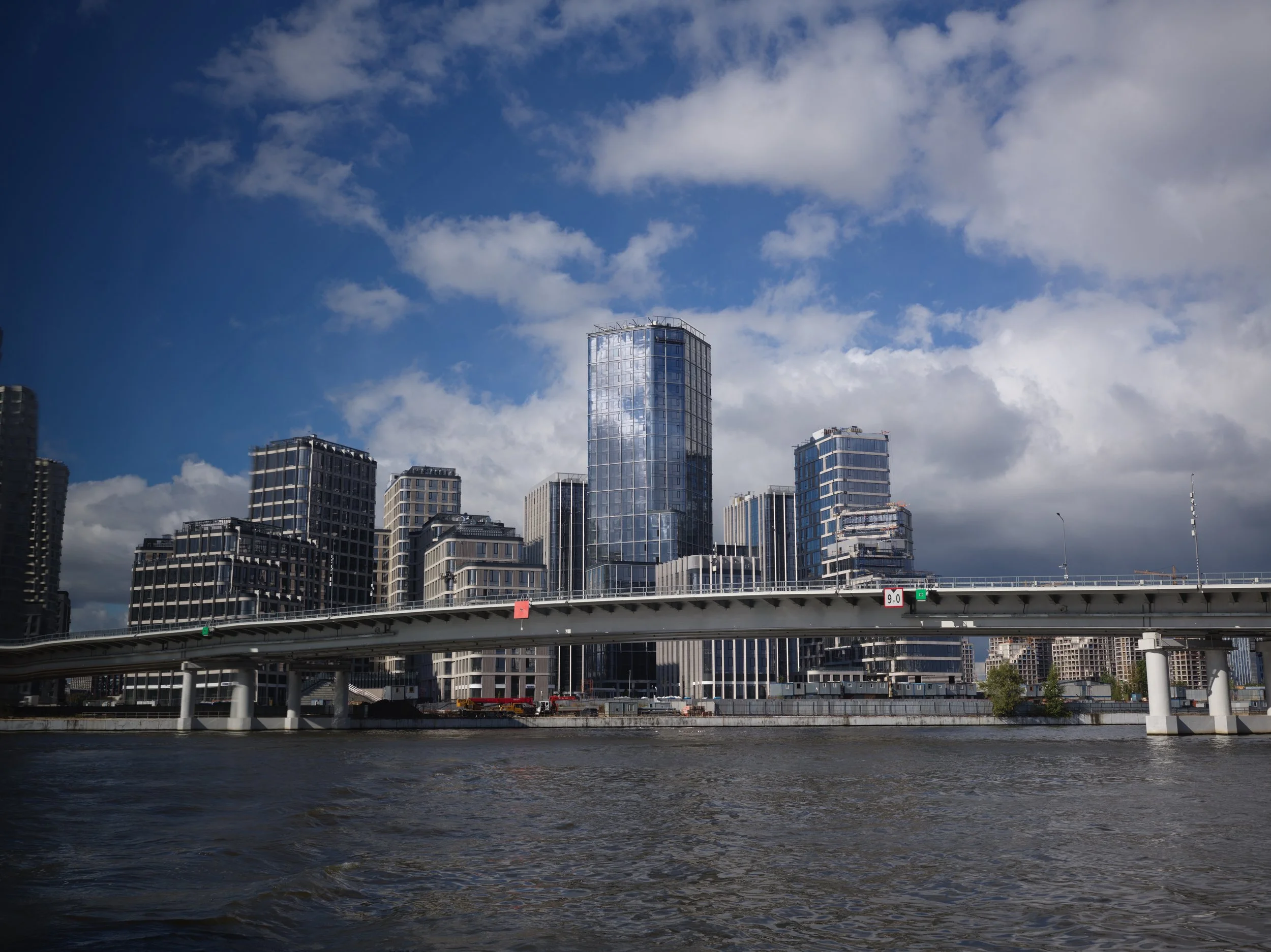 City skyline with modern glass skyscrapers along a river under a partly cloudy sky.