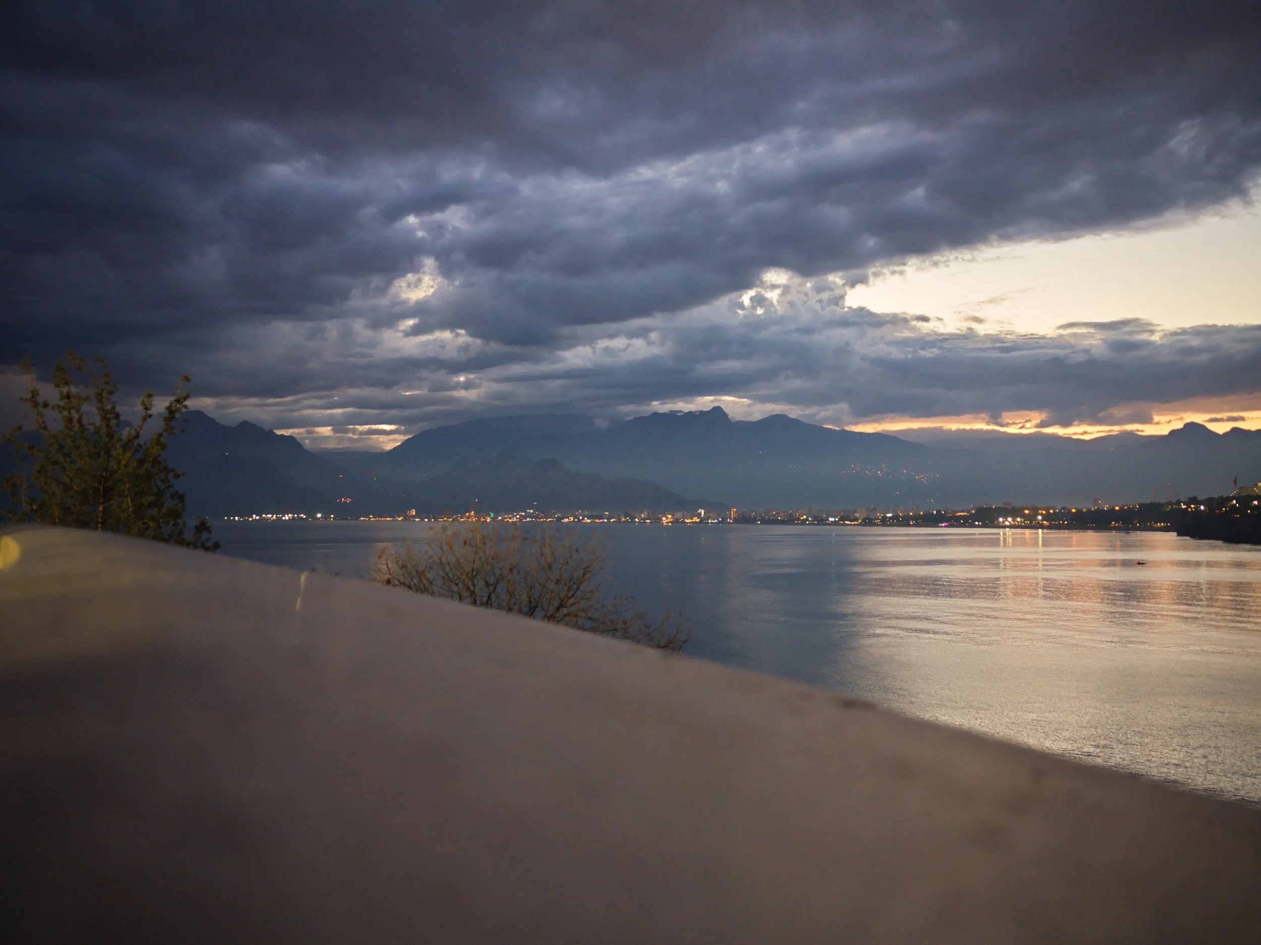 A view of a river at dusk with mountains in the background and city lights along the shore, cloudy sky overhead.