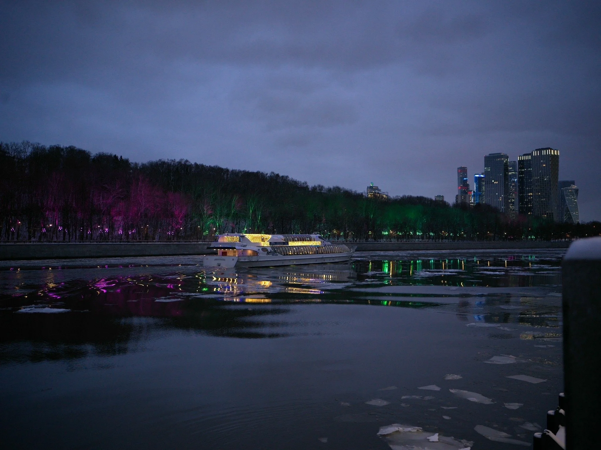 Nighttime cityscape with high-rise buildings, a boat on a river, and illuminated trees along the riverbank. Photo by Alexander Sokolyuk. Moscow, Russia