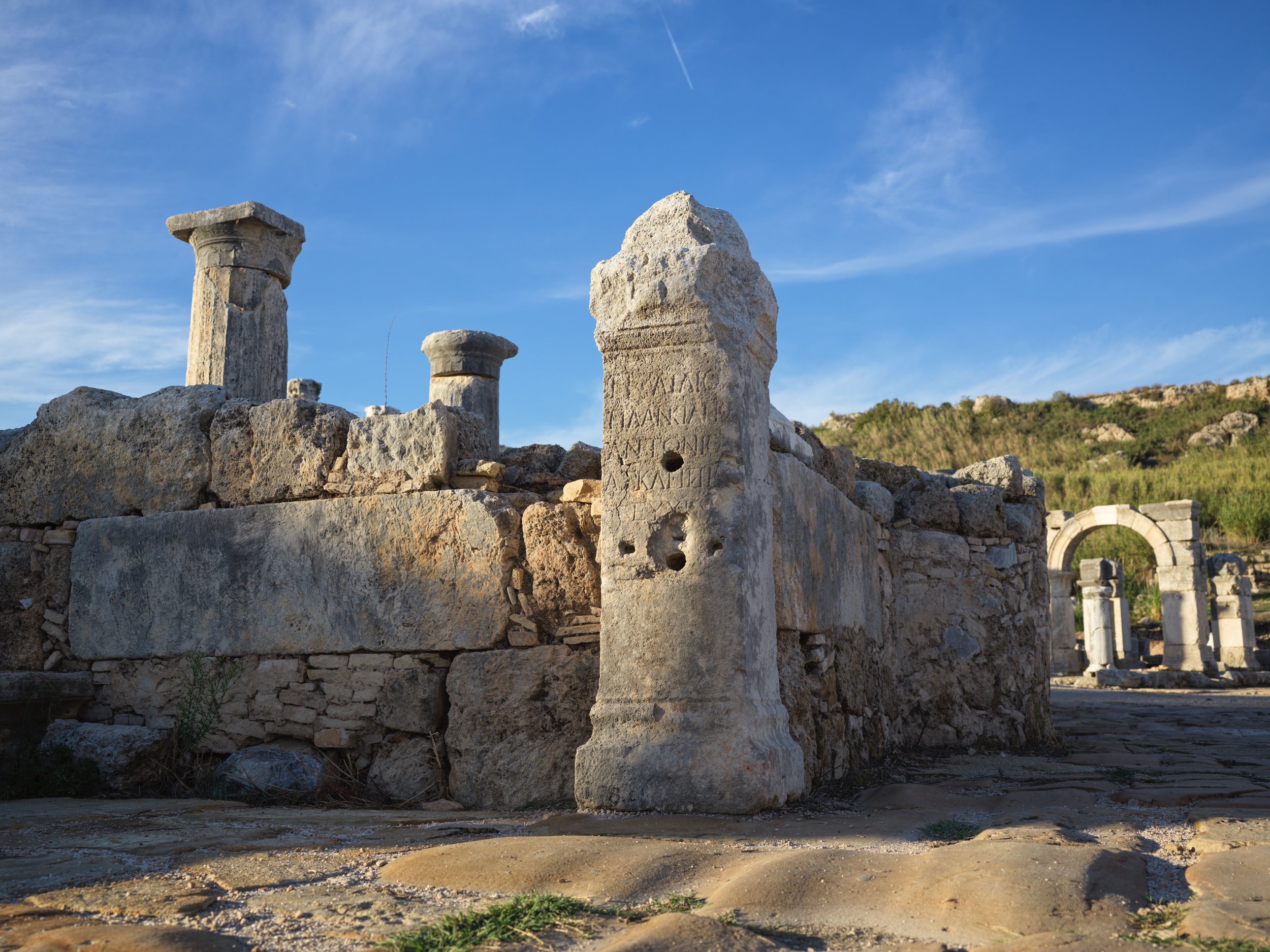 Ancient ruins with stone columns and arch structures under a clear blue sky, with a hillside in the background.