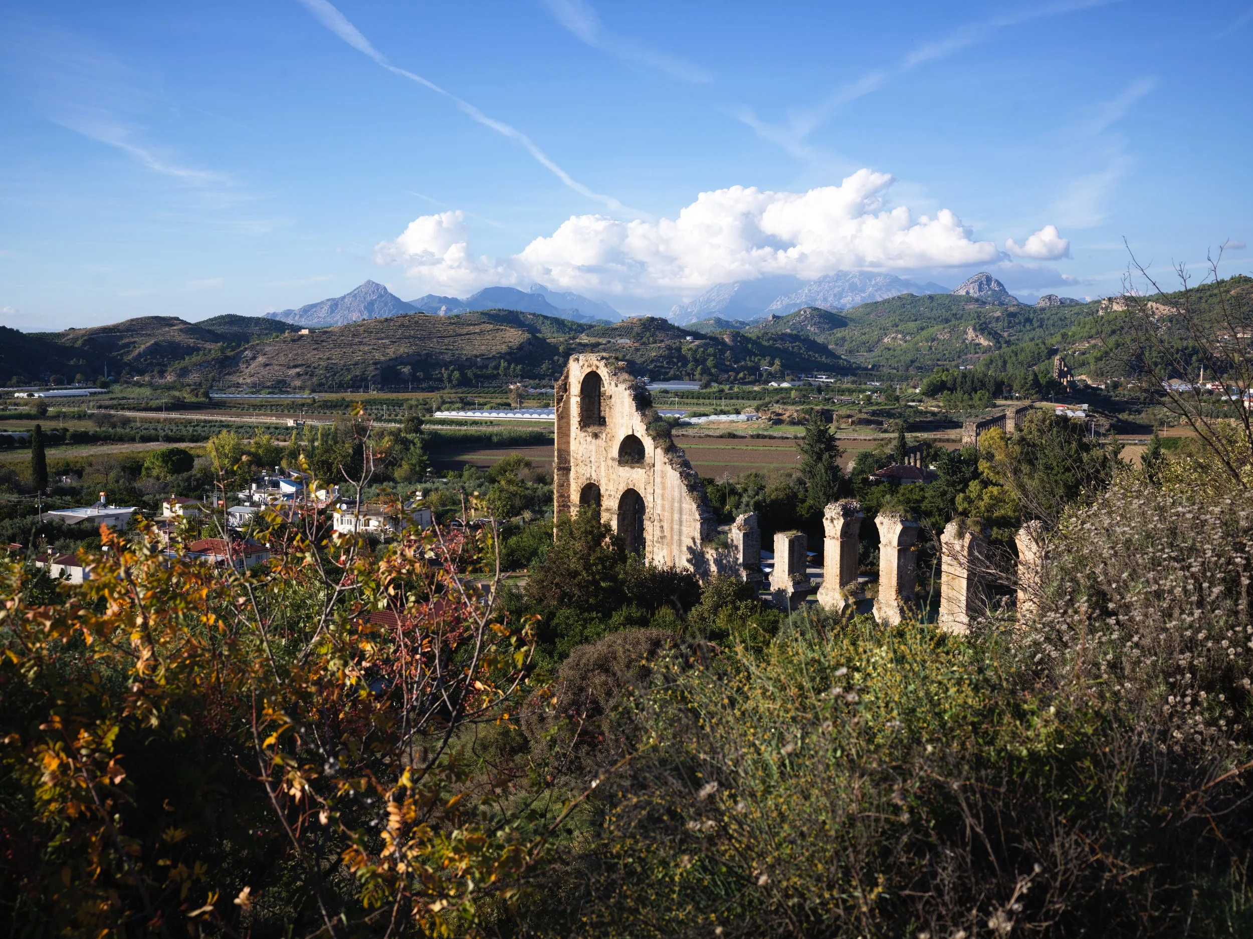 Scenic landscape with ancient ruins, green fields, trees, mountainous backdrop, and a partly cloudy sky.