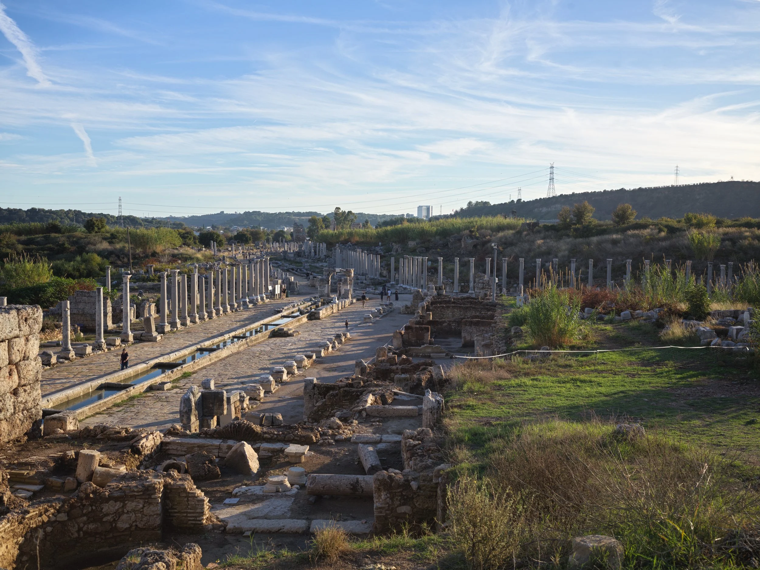 Ancient ruins with stone columns and pathways, surrounded by greenery and hills under a partly cloudy sky.