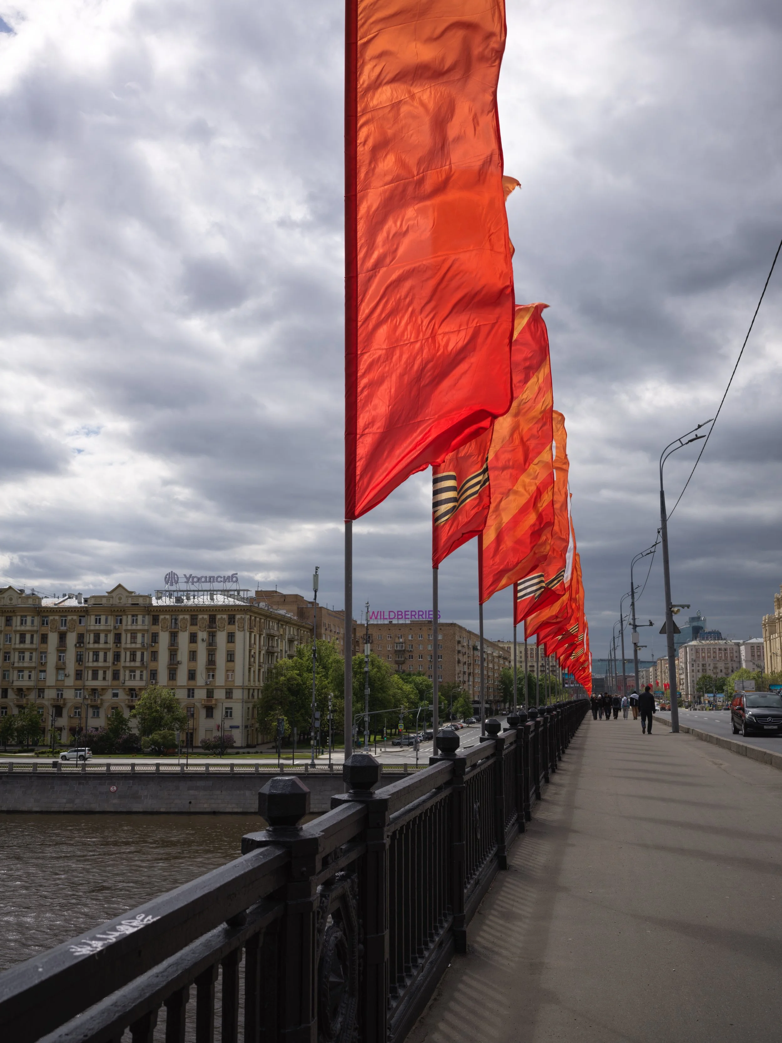Red flags line a bridge with gray clouds overhead and city buildings in the background.