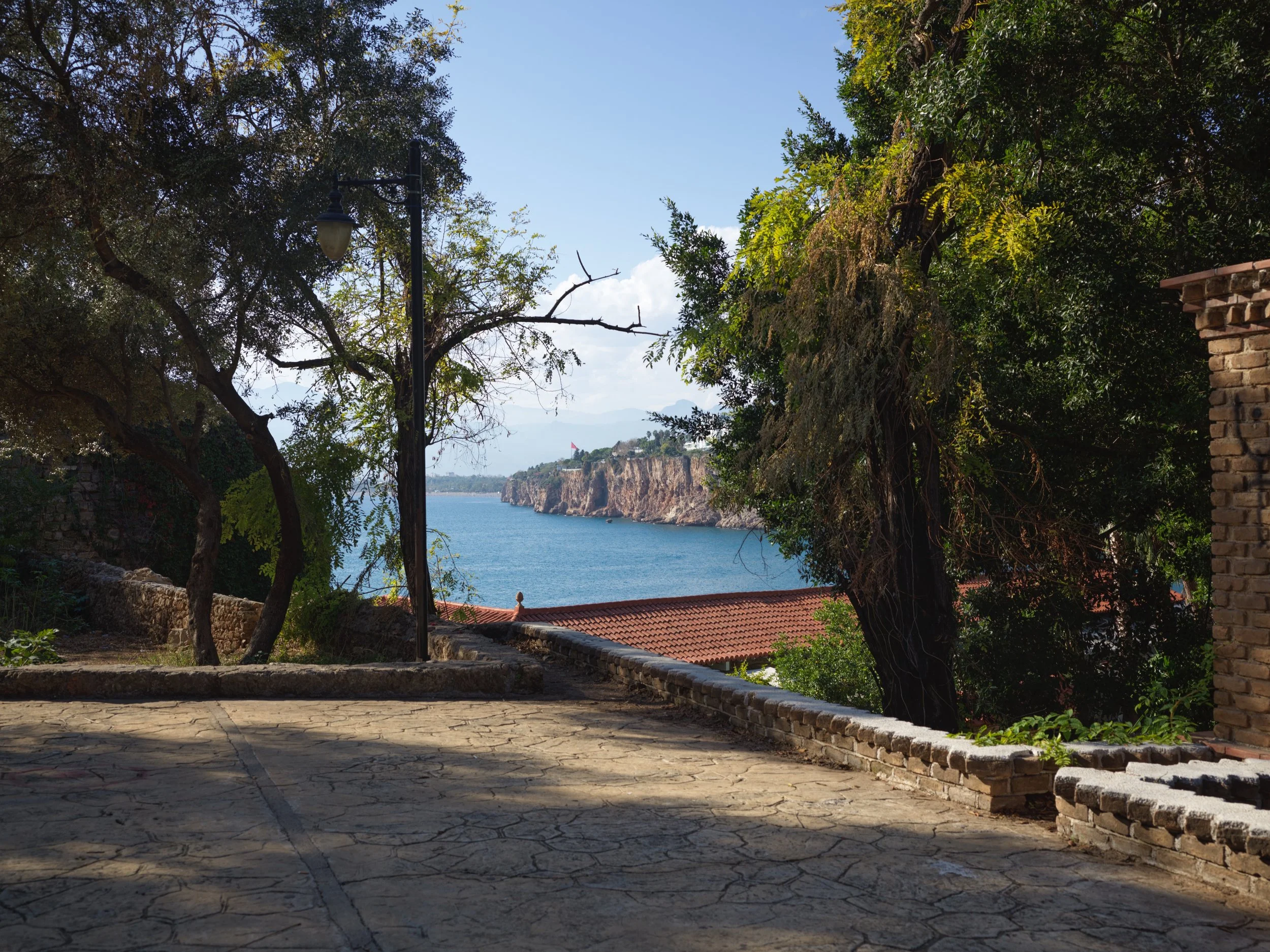 Paved outdoor area with a tree and a street lamp, overlooking a body of water and rocky cliffs in the distance under a partly cloudy sky.