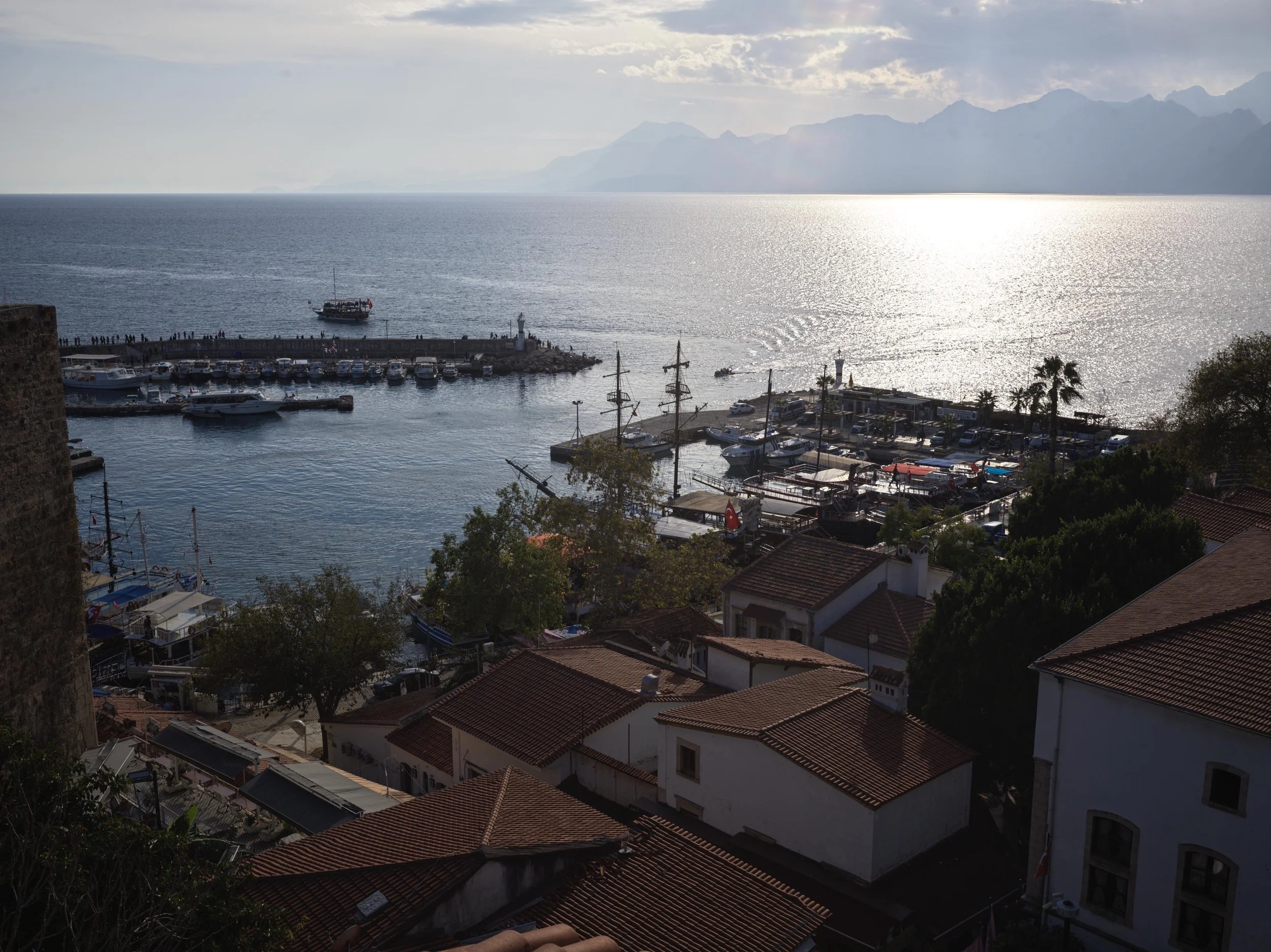 A scenic view of a harbor with multiple boats, a pier, and a ship in the water. There are houses with red-tile roofs in the foreground, trees, and distant mountains under a cloudy sky with sunlight reflecting on the water.