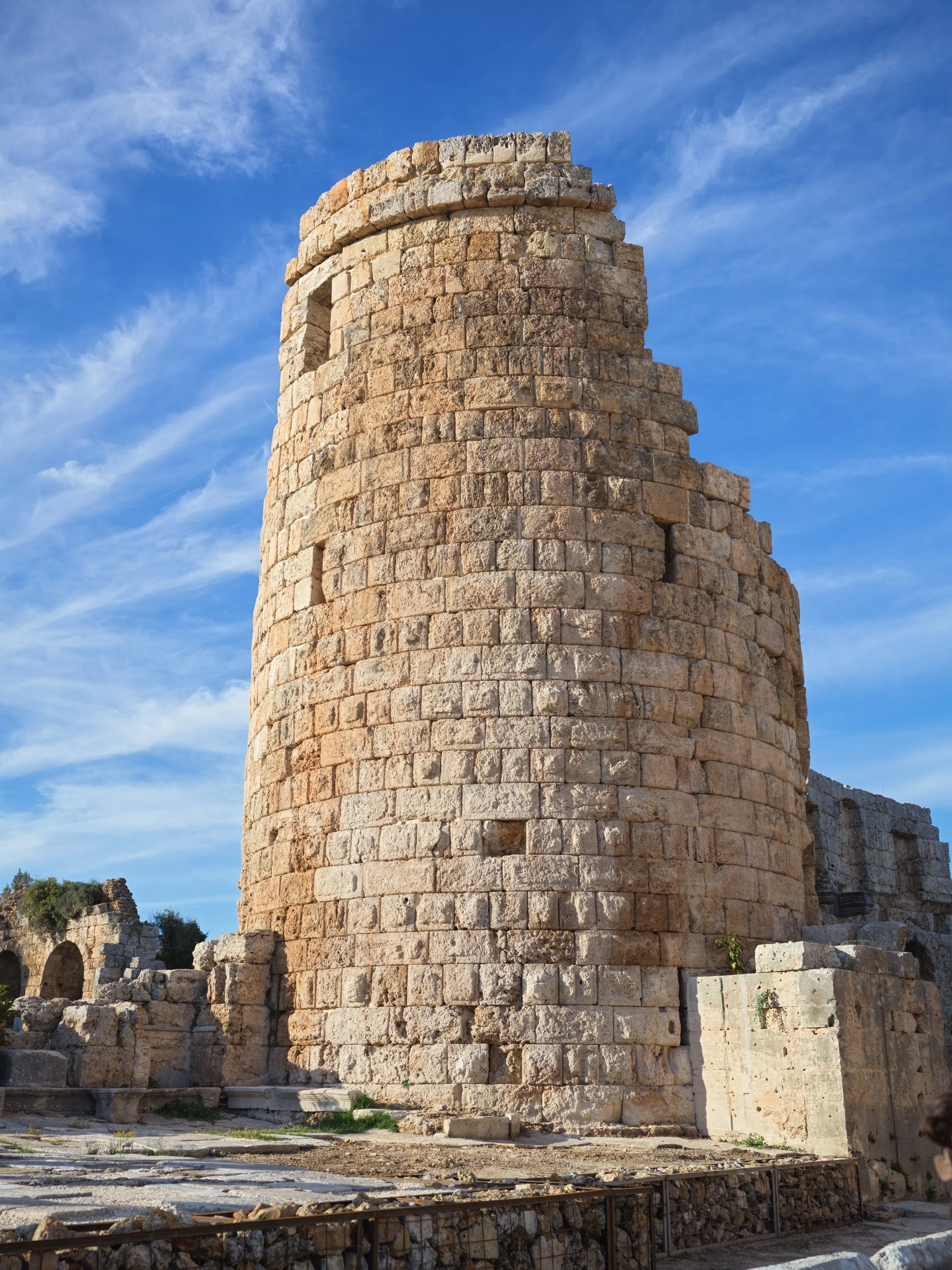 Ancient stone tower with a cylindrical shape, partially reconstructed, situated under a clear blue sky with wispy clouds. Photo by Alexander Sokolyuk. Old ancient city PERGE in Türkiye
