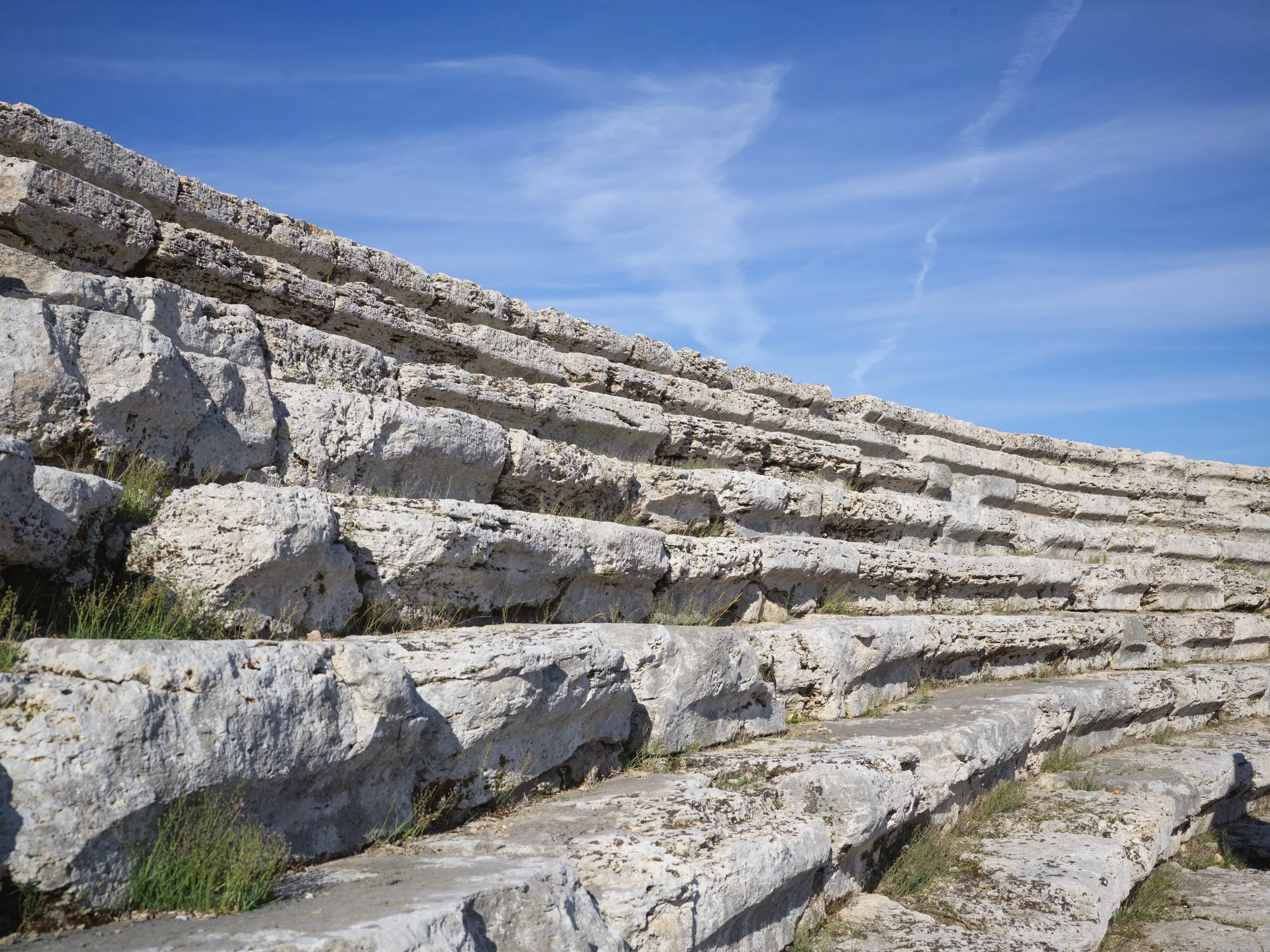 Ancient stone amphitheater with tiered seating under a blue sky with a few wispy clouds.