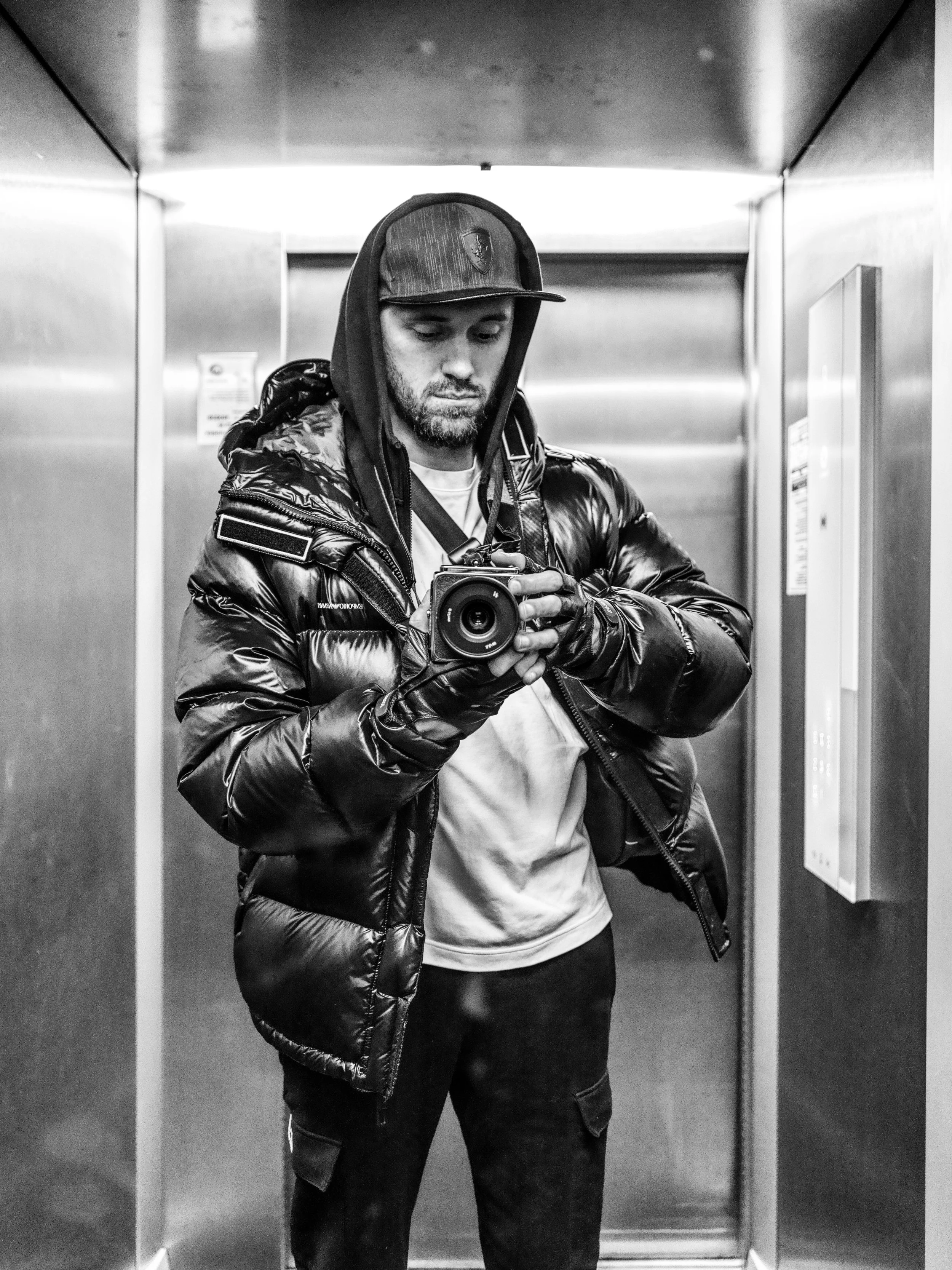 A man taking a selfie in an elevator, dressed in a shiny black puffer jacket and a baseball cap.