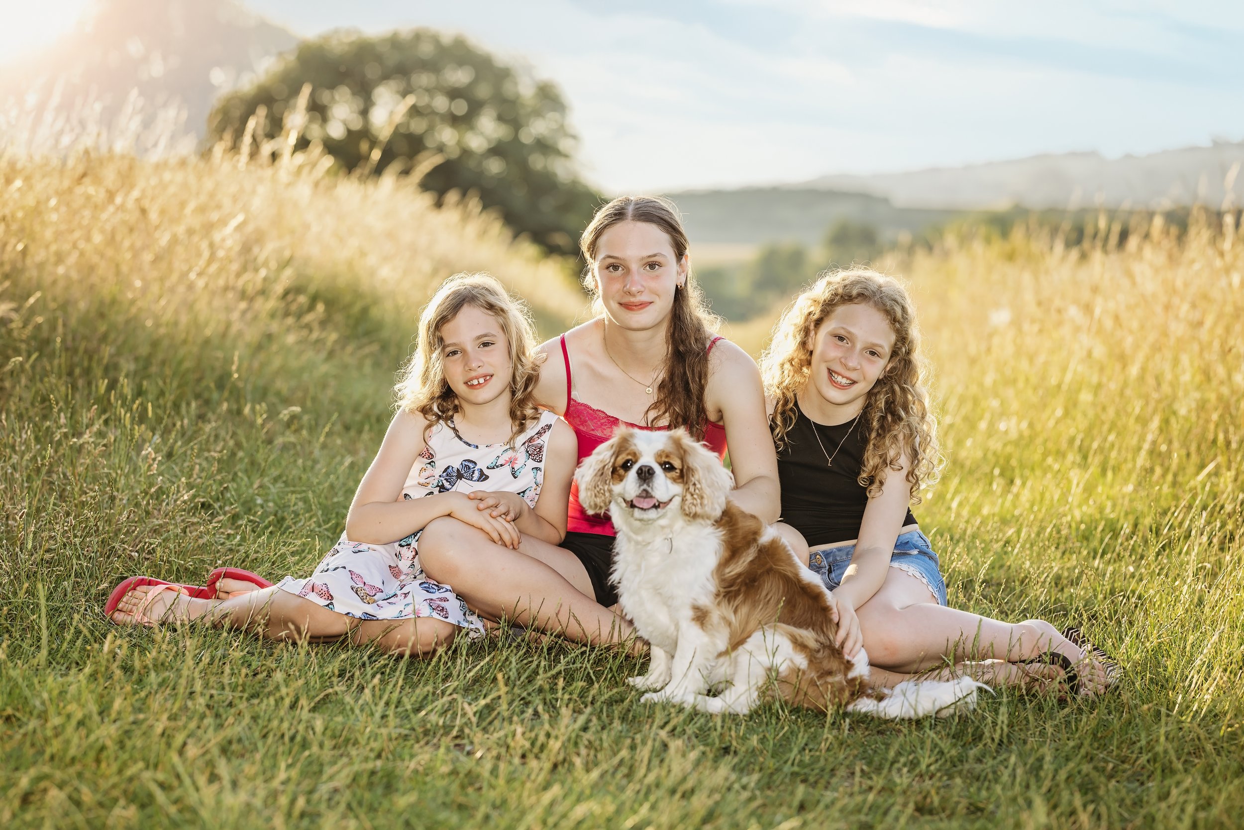 Three girls sitting on grass in a field with a dog during sunset, smiling at the camera.