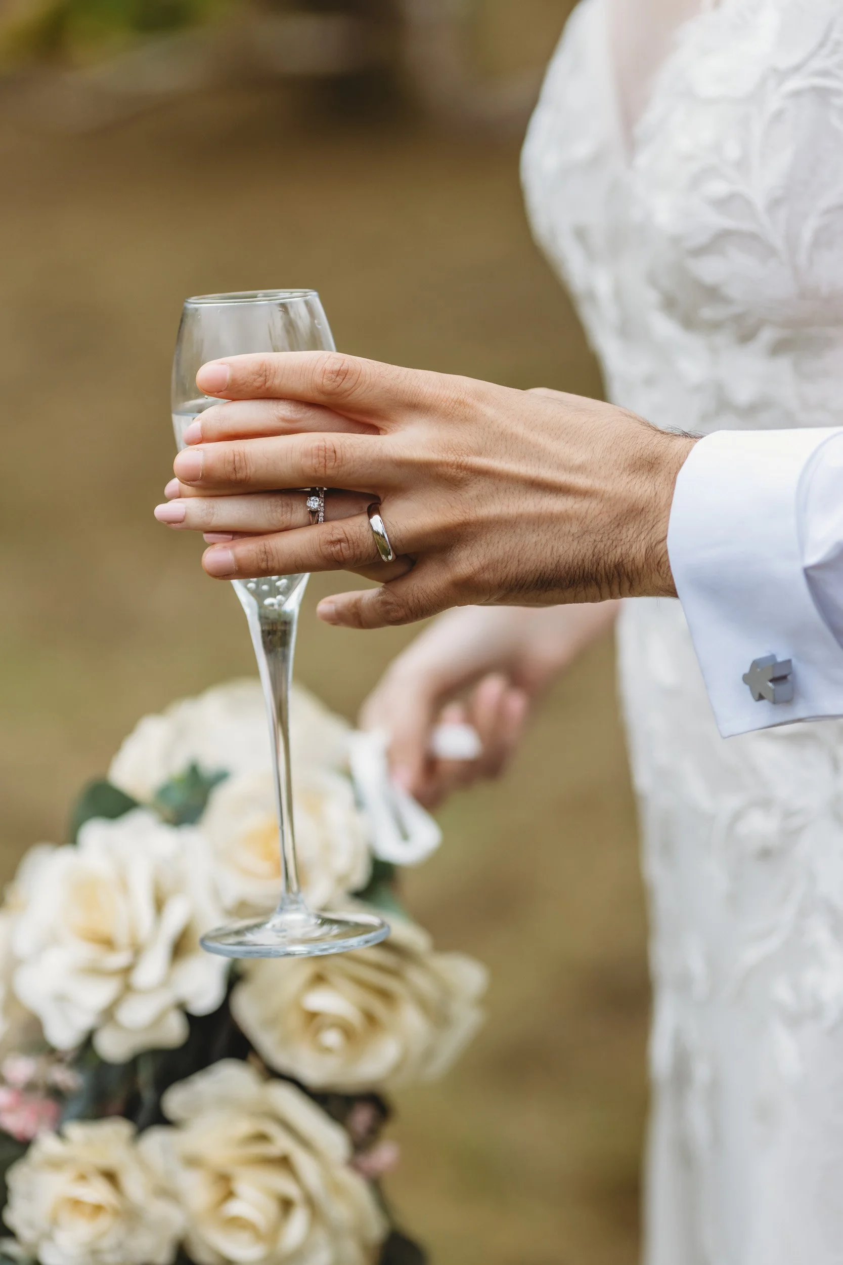 A person holding a glass of champagne with wedding rings on their finger, wearing a white wedding dress, with a bouquet of white roses in the background.