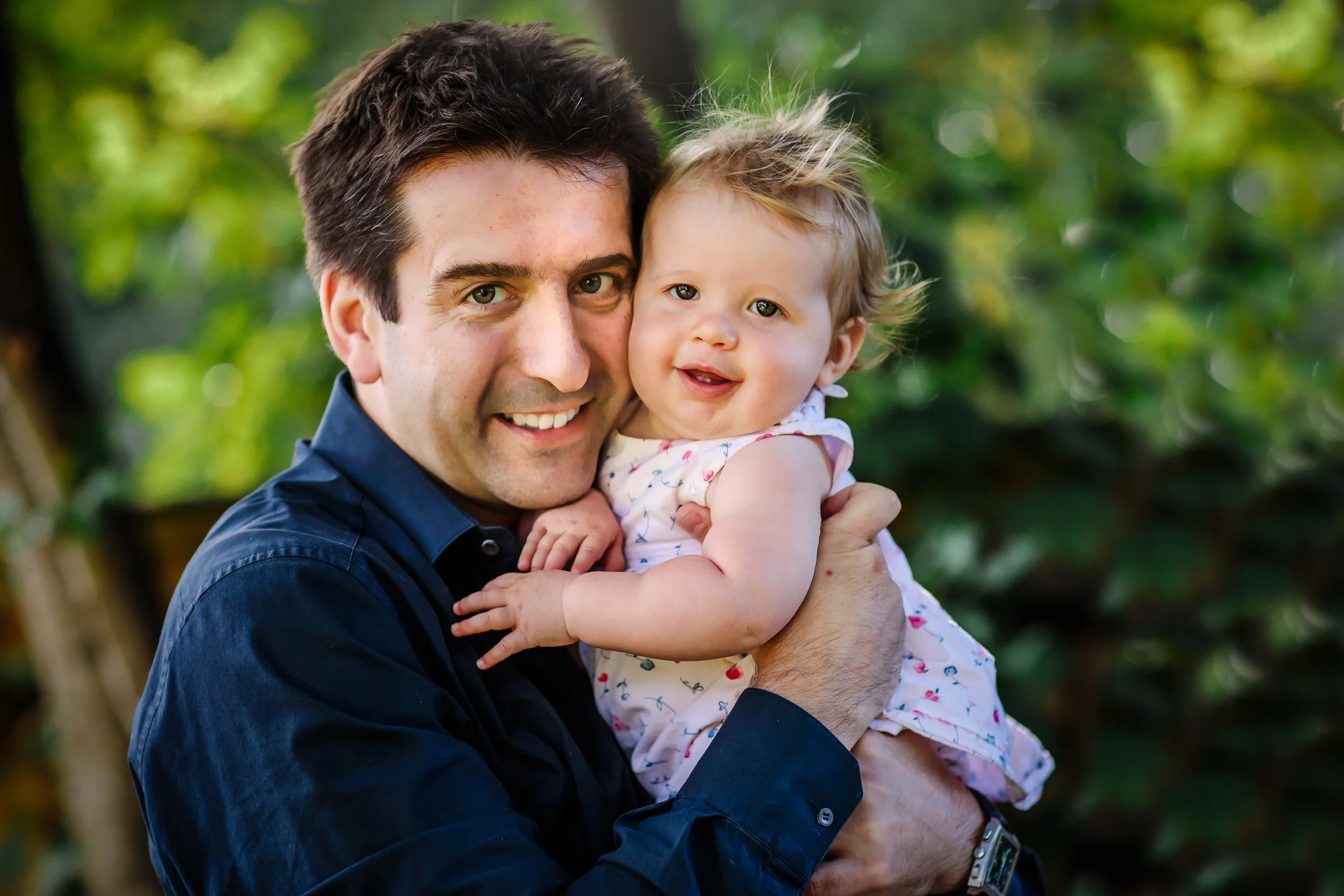 A man holding a smiling young girl outdoors in a green, leafy area.