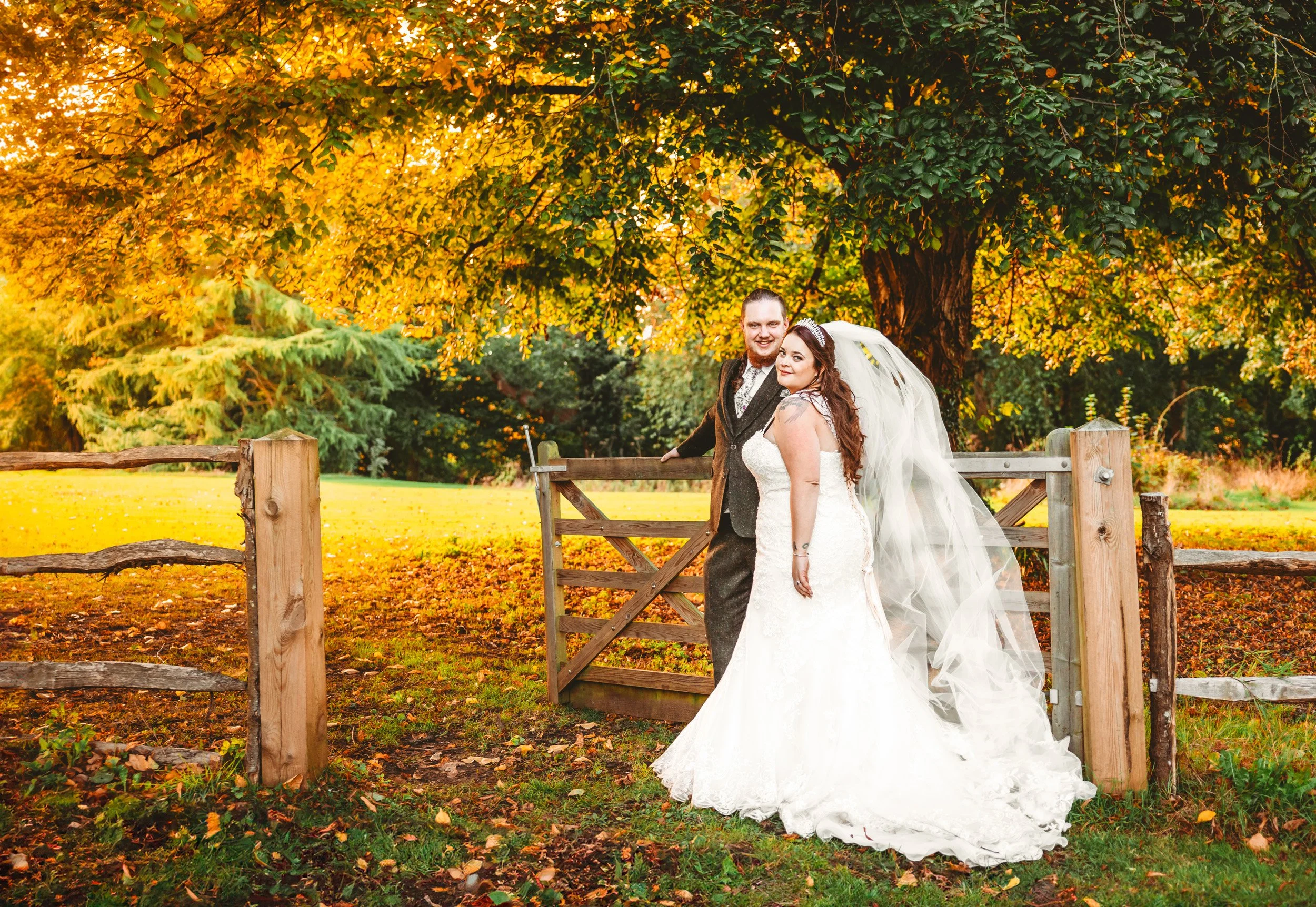 A newlywed couple in wedding attire standing in front of a wooden gate with a large tree with orange and yellow autumn leaves in the background.