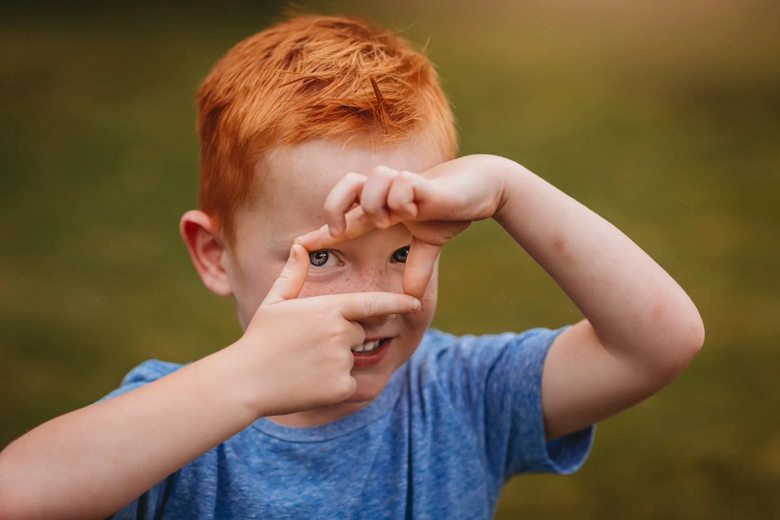 A young red-haired boy with freckles wearing a blue shirt is playfully making a frame with his hands around his eye, looking through it with a smile.
