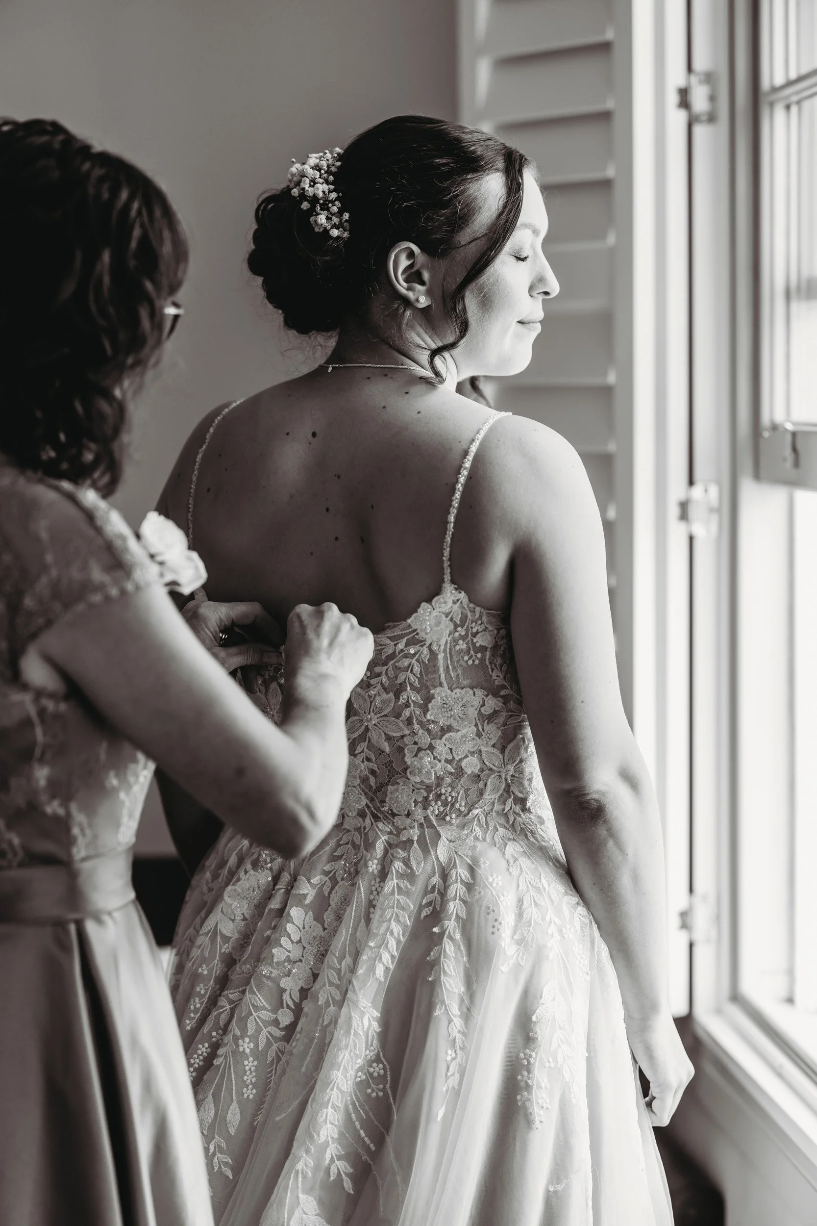 A bride in a lace wedding dress standing by a window, smiling with eyes closed, as another woman pinches the back of her dress.