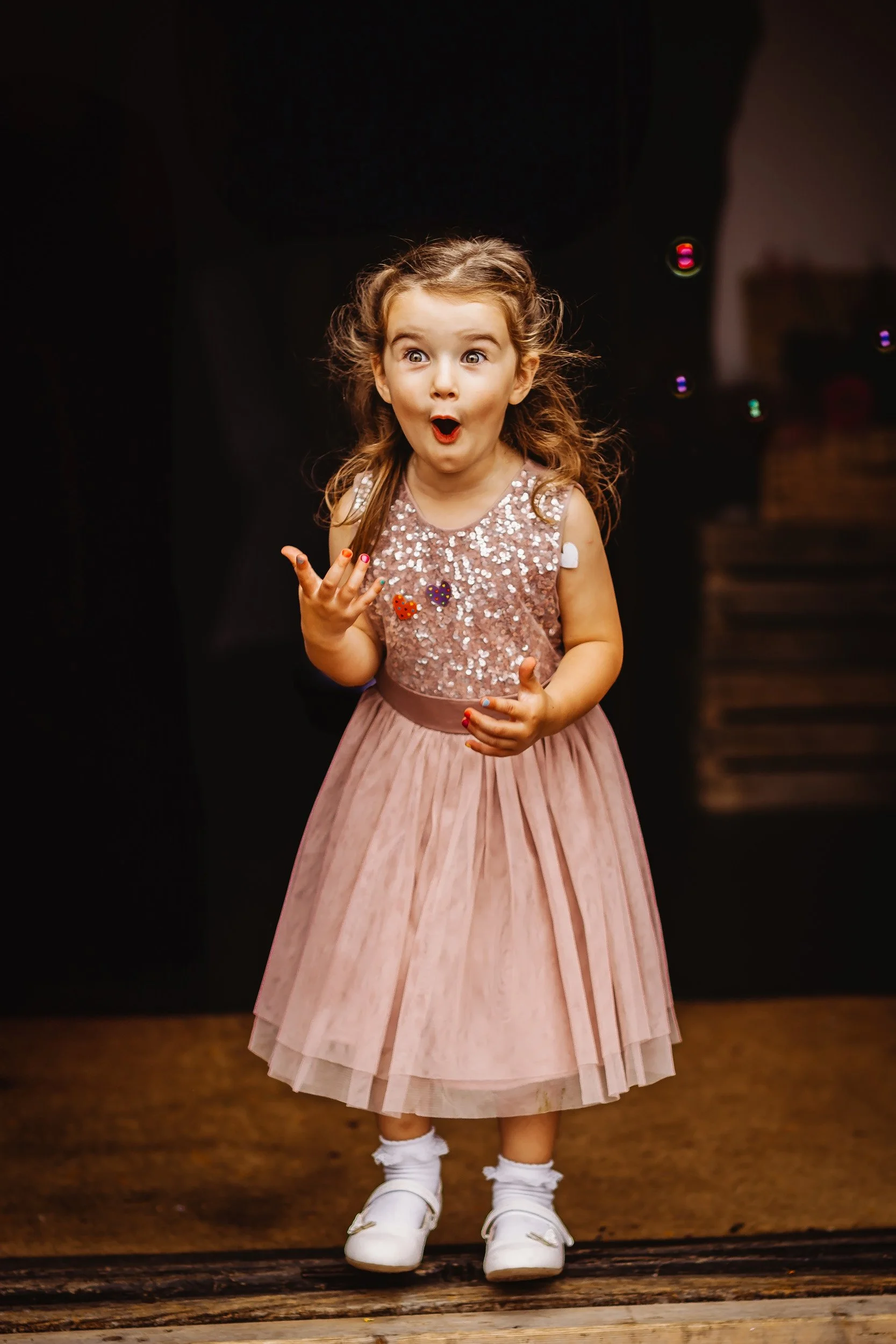 A young girl with curly hair, dressed in a sparkly pink dress and white shoes, has an amazed or surprised expression on her face, with her mouth open and her hand raised.