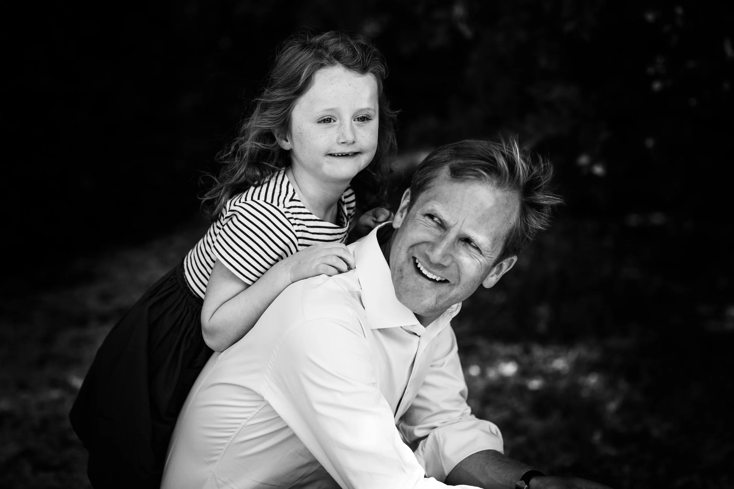 A man and a young girl sharing a joyful moment outdoors, with the girl sitting on the man's back and both smiling.