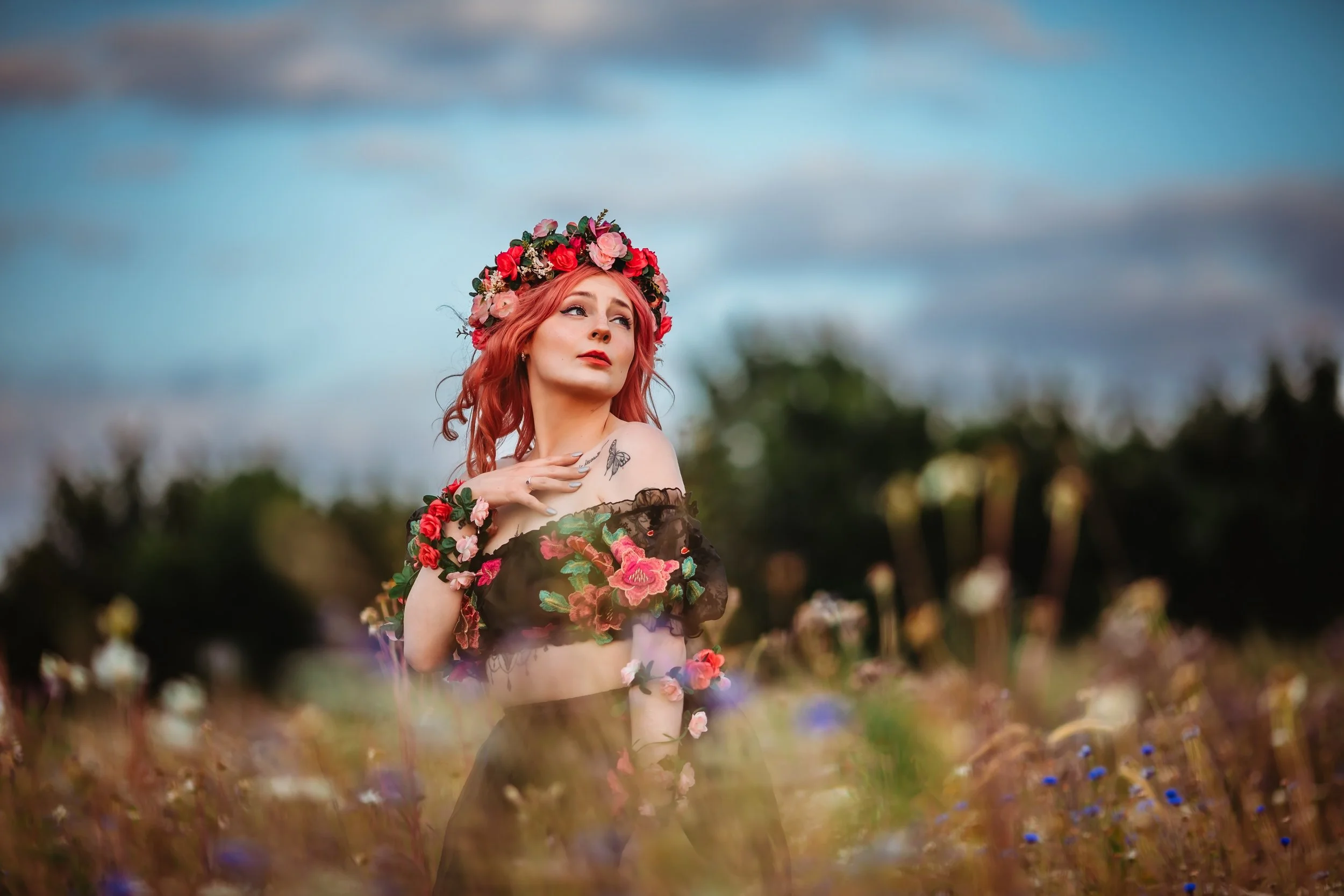 A woman with pink hair wearing a flower crown and a black off-the-shoulder dress with floral embroidery, standing in a field of wildflowers under a cloudy sky.