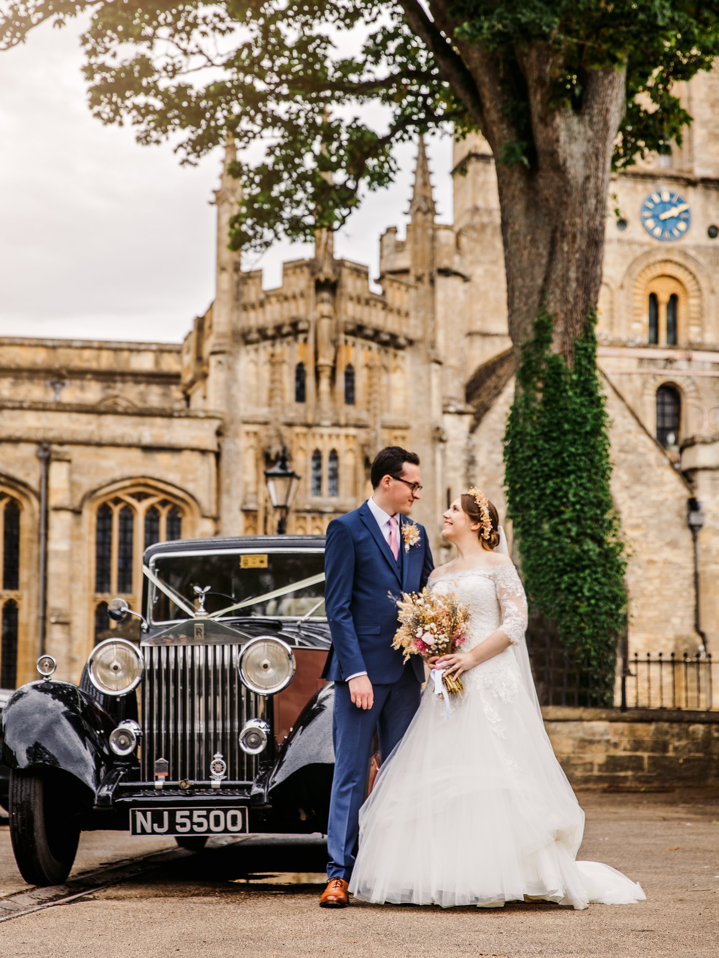 A bride and groom in wedding attire standing beside a vintage black car with a church in the background, holding a flower bouquet.