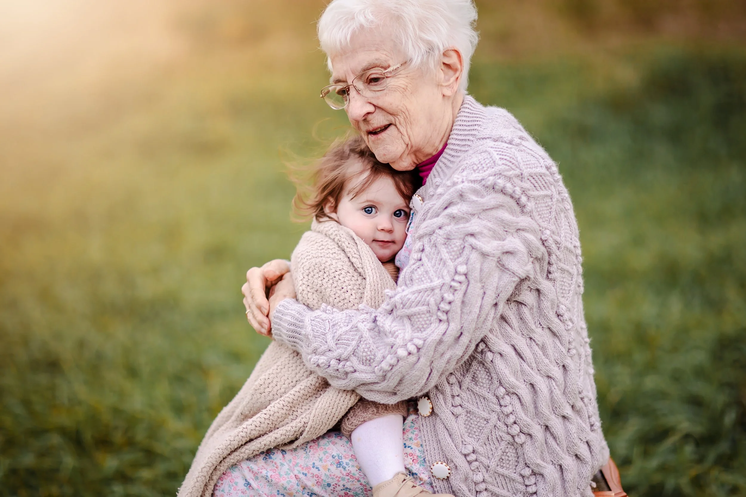 An elderly woman holding a young girl, both wearing cozy knit sweaters, outdoors in a grassy area with a blurred background.