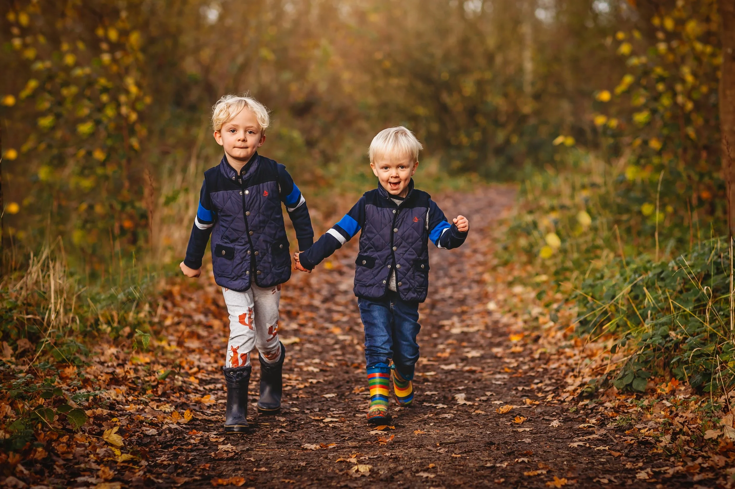 Two young children, a boy and a girl, holding hands and running along a dirt path in a forest during fall, surrounded by colorful autumn leaves.