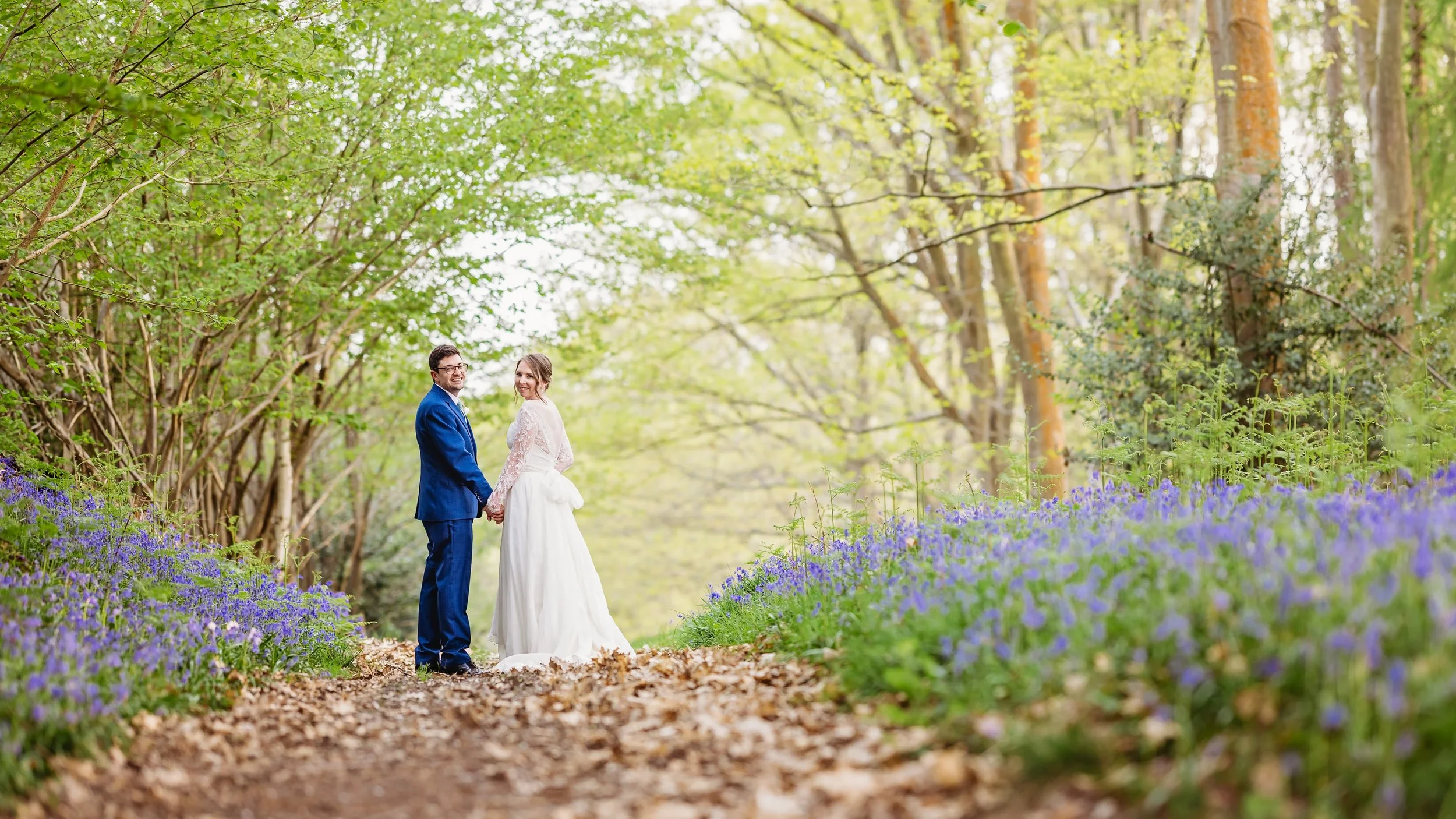 A newlywed couple holding hands and smiling while standing on a wooded trail covered with fallen leaves, surrounded by purple wildflowers and green trees in springtime.