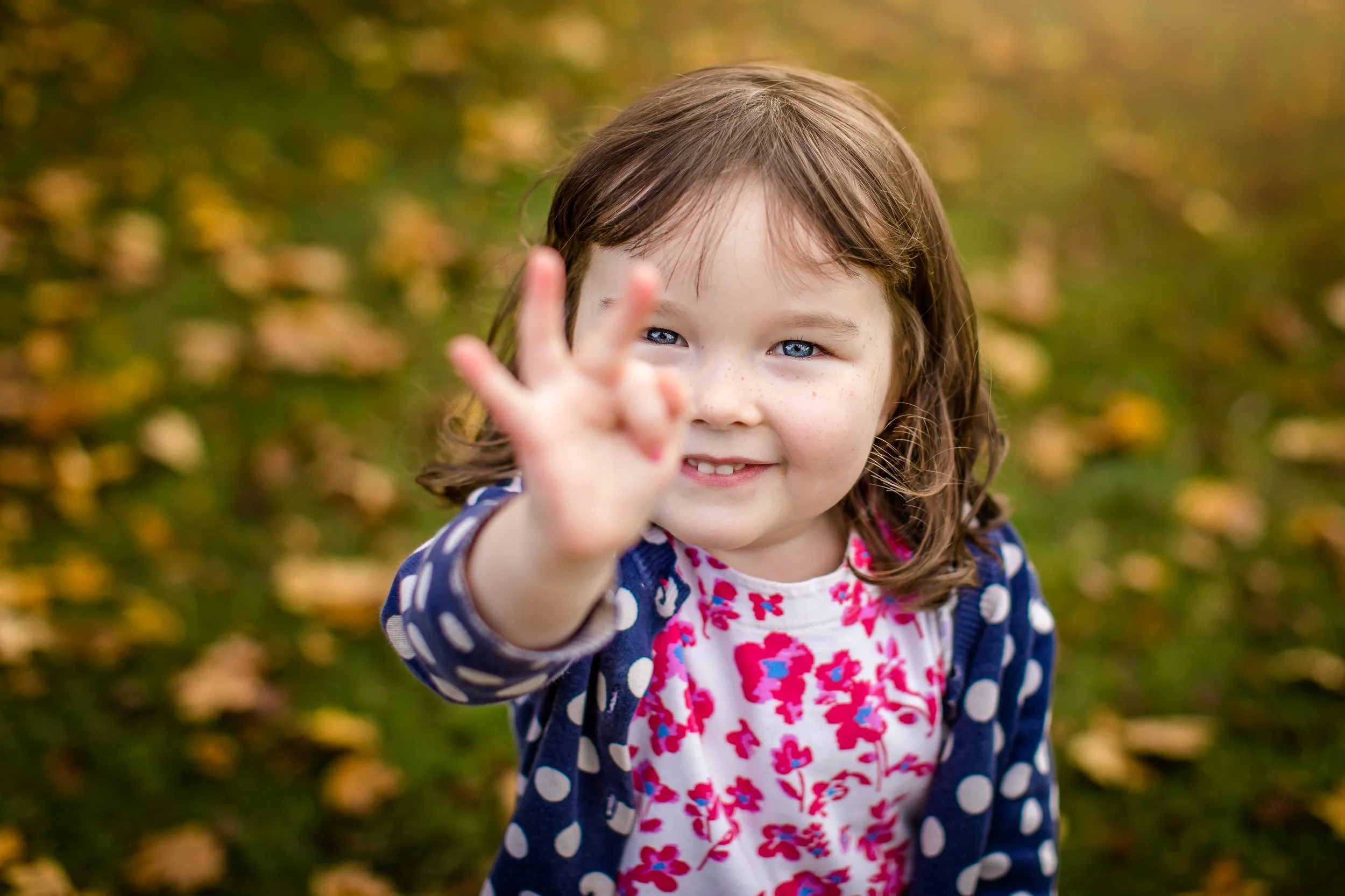 A young girl with brown hair, blue eyes, and freckles, smiling and reaching out her hand in a peace sign gesture during fall with fallen leaves in the background.
