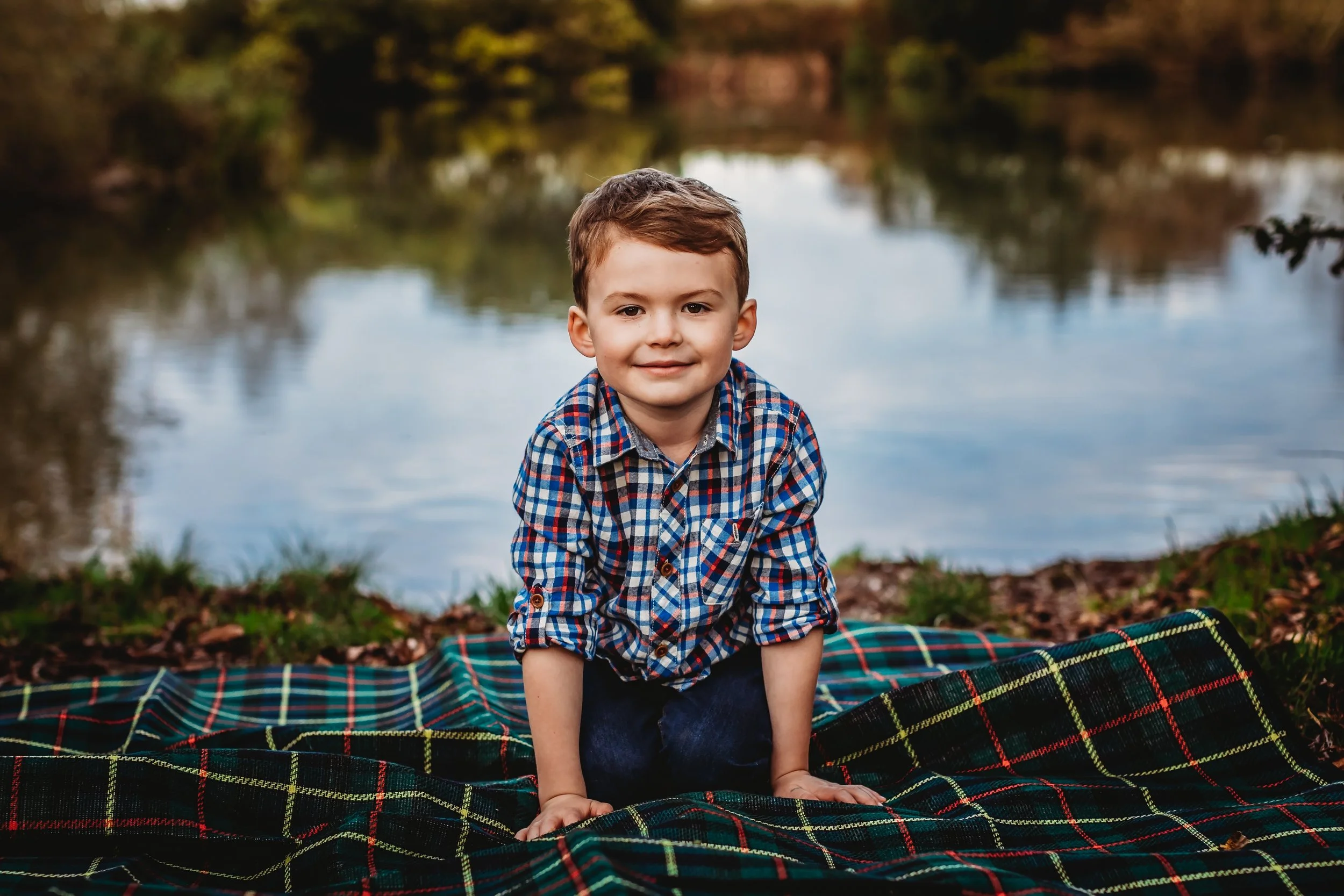 A young boy with short brown hair, wearing a plaid shirt and jeans, is kneeling on a plaid blanket by a lake, with trees and water in the background.