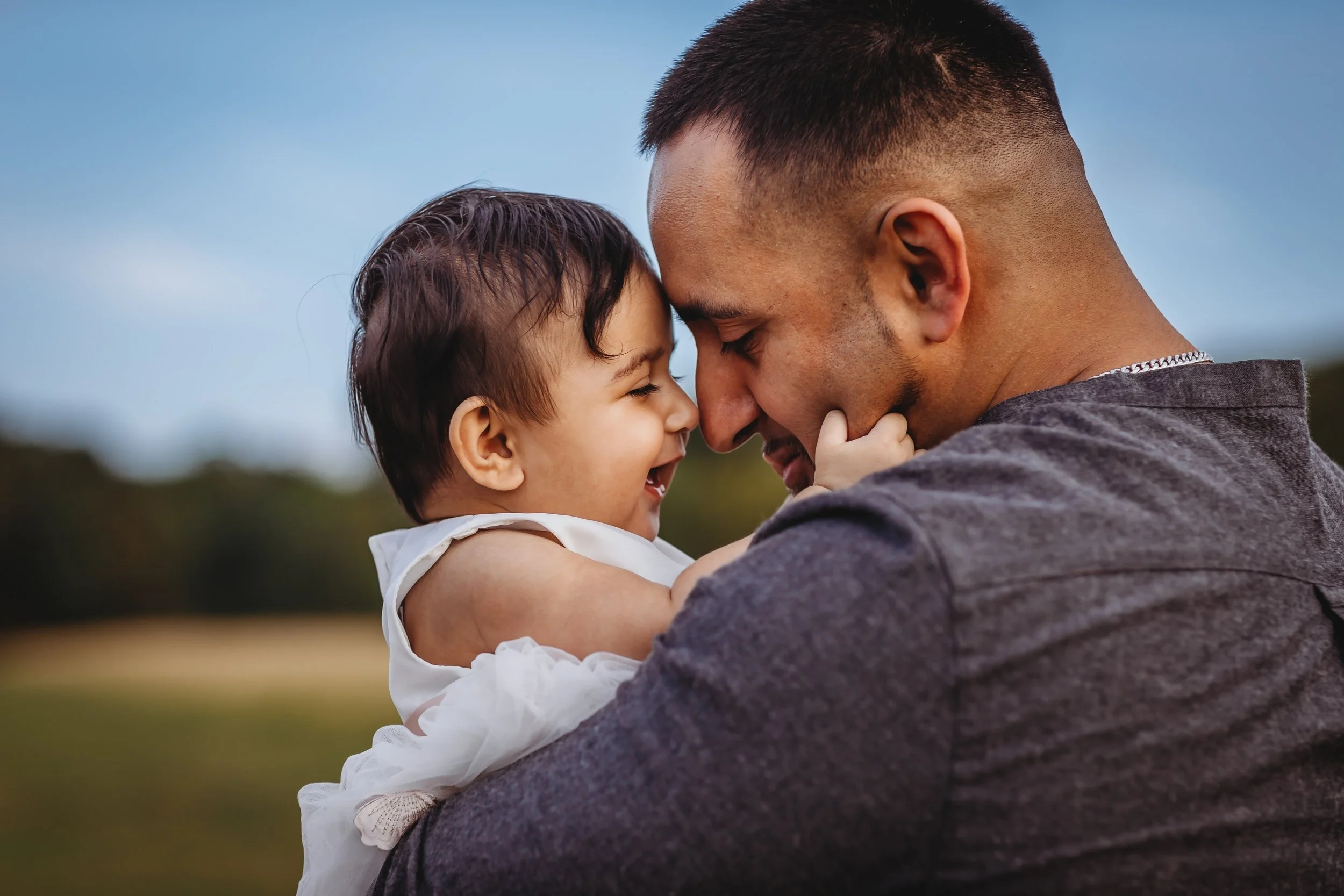 A close-up of a father and his young daughter touching foreheads and smiling together outdoors with a blurred background of trees and sky.