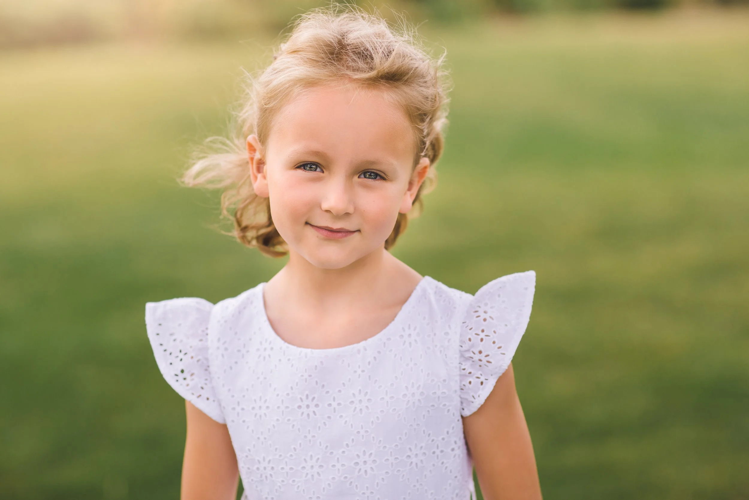 A young girl with blonde, curly hair and blue eyes, wearing a white eyelet dress, standing outdoors with a blurred grassy background.