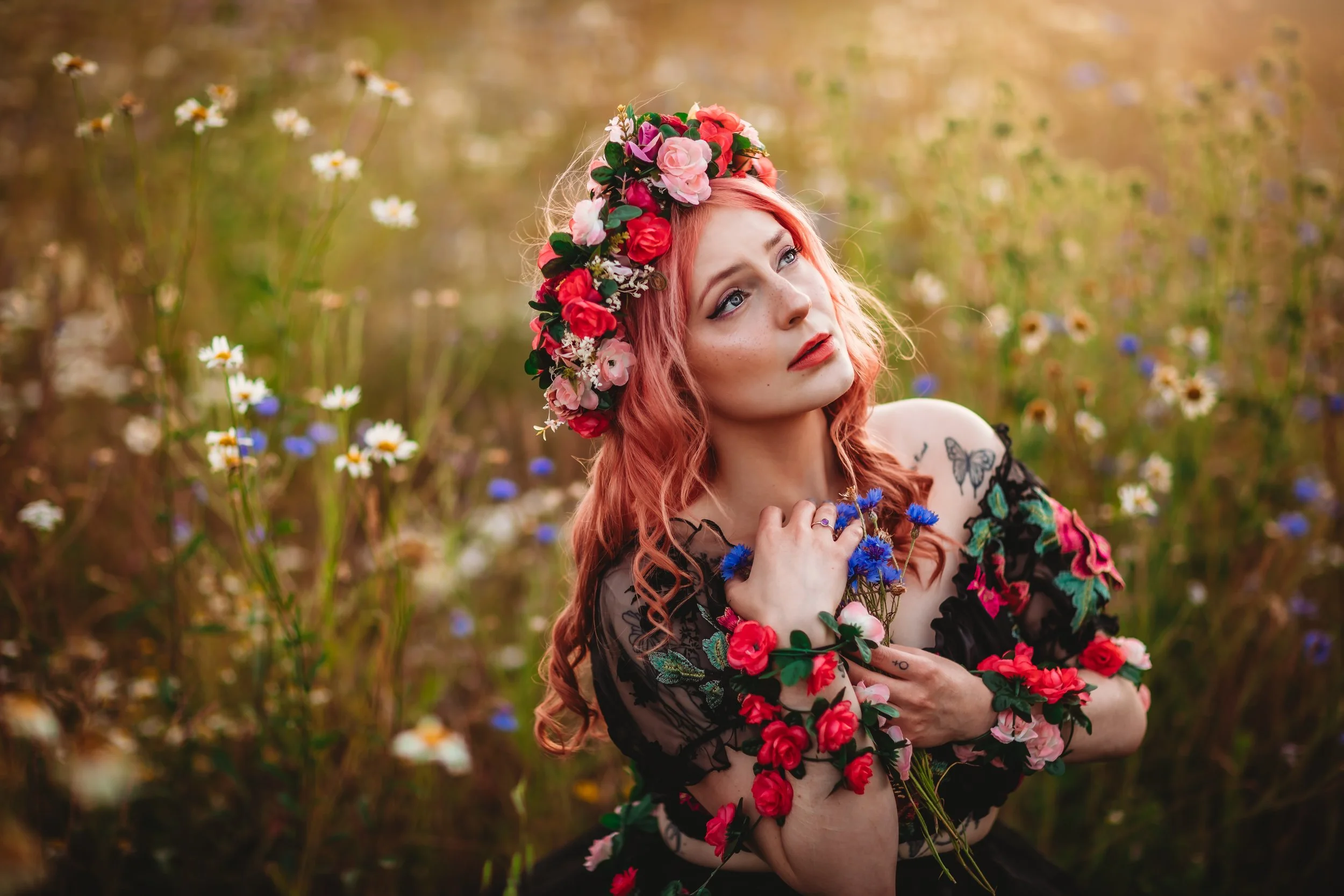 A woman with pink hair in a floral crown and floral accessories on her black dress is sitting in a field of wildflowers, including daisies, cornflowers, and other colorful blooms, with a thoughtful expression.