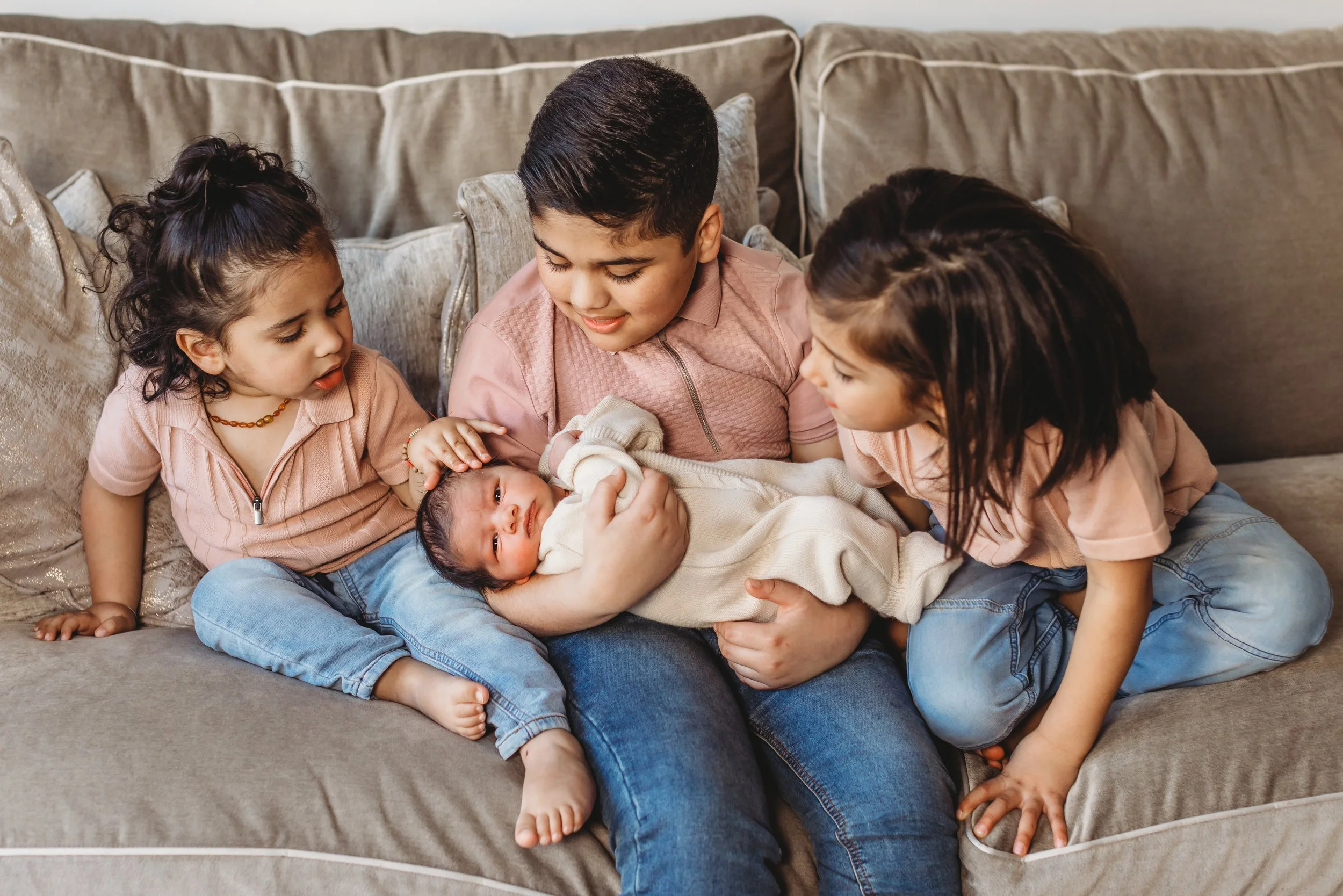 A boy and three young girls sitting on a beige couch, holding and looking at a newborn baby.