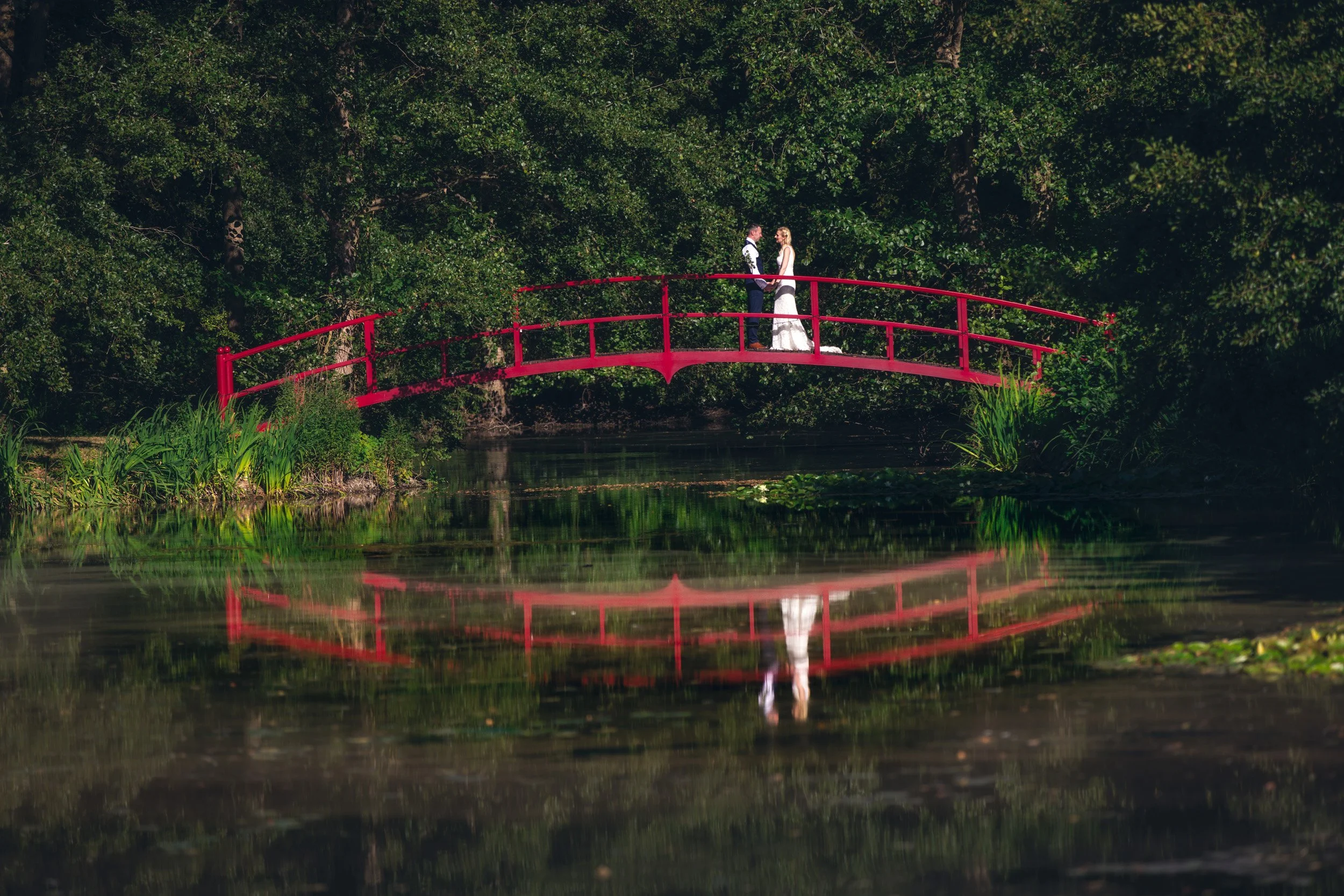 A bride and groom stand on a small red arched bridge over a calm river, surrounded by lush green trees and plants.