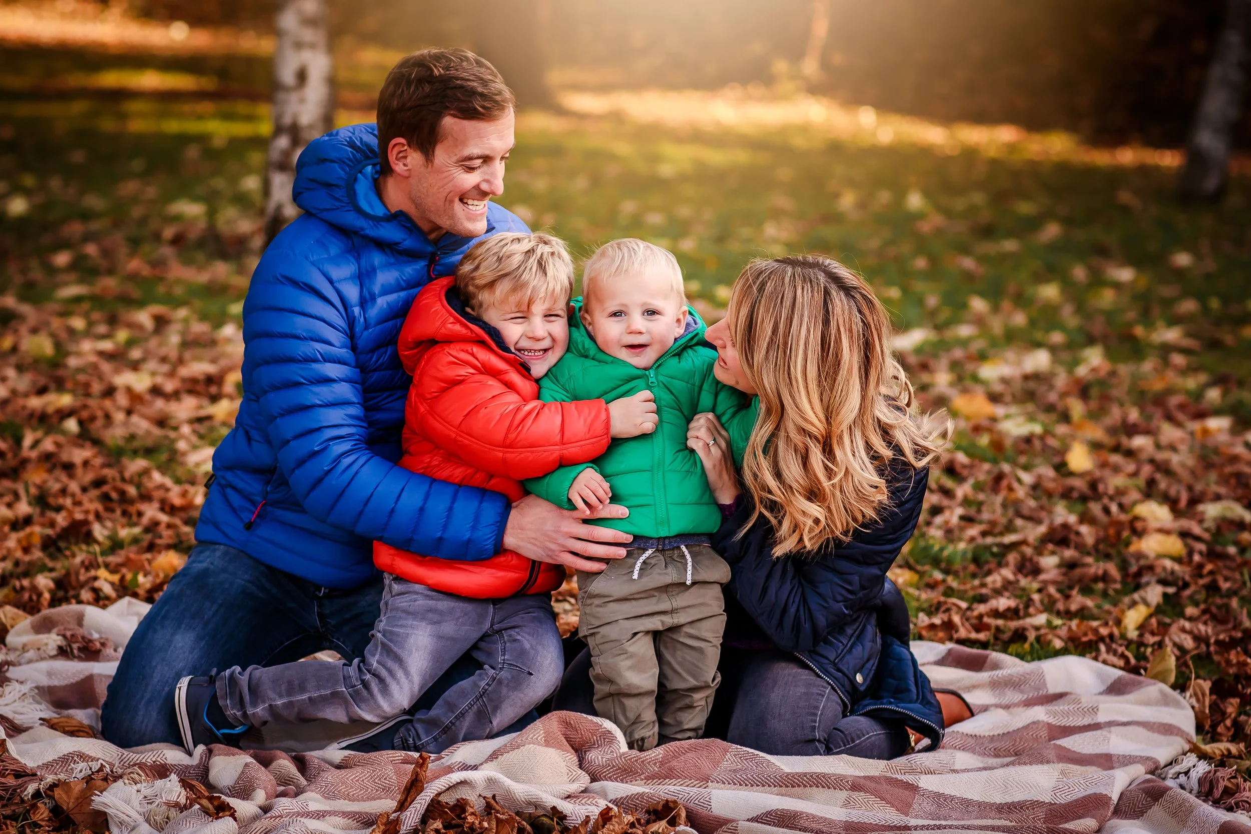 A family of four in autumn clothing sitting on a blanket in a park surrounded by fallen leaves, smiling and playing together.