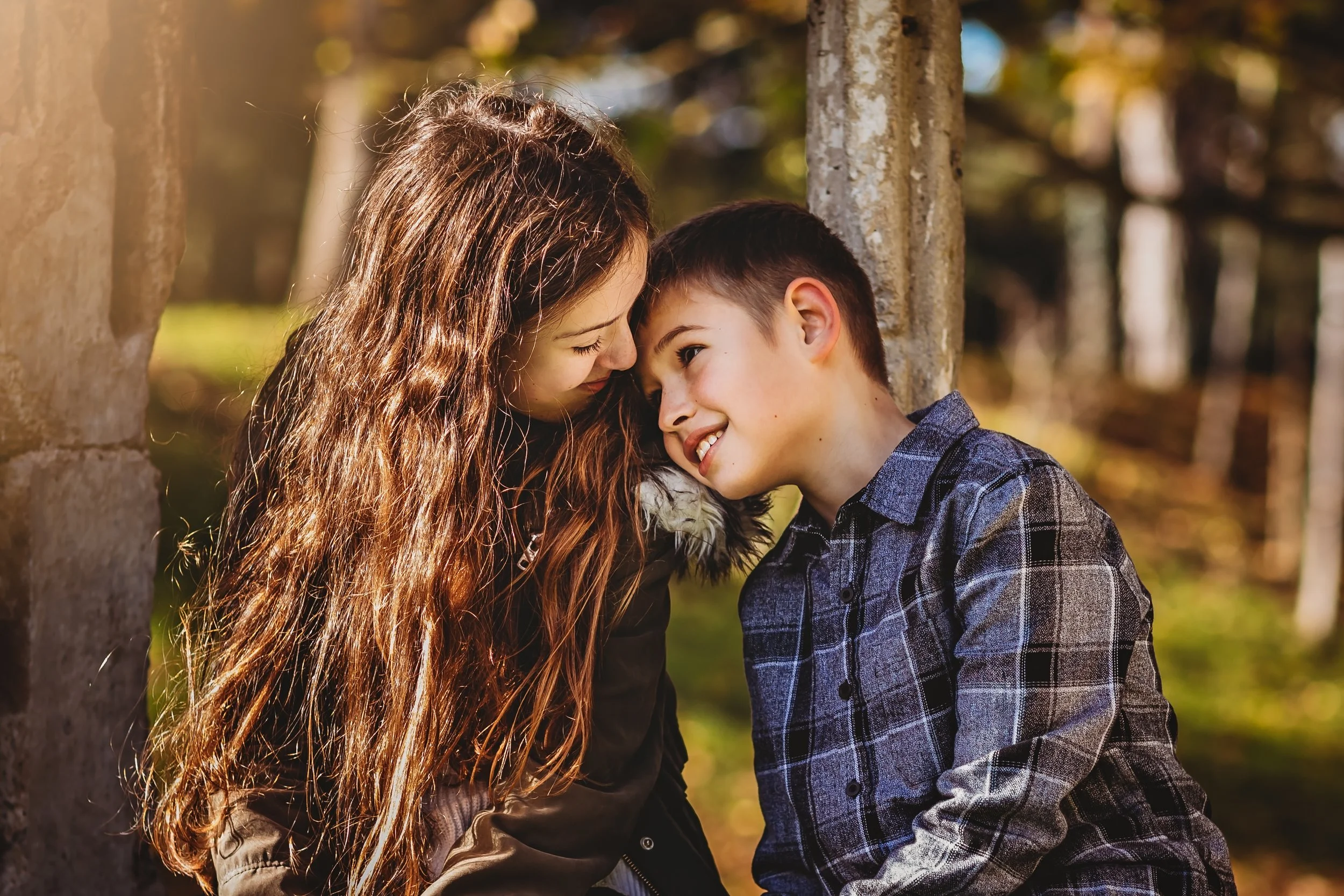 A young girl and boy lean their foreheads together, smiling in an outdoor park with trees and sunlight in the background.