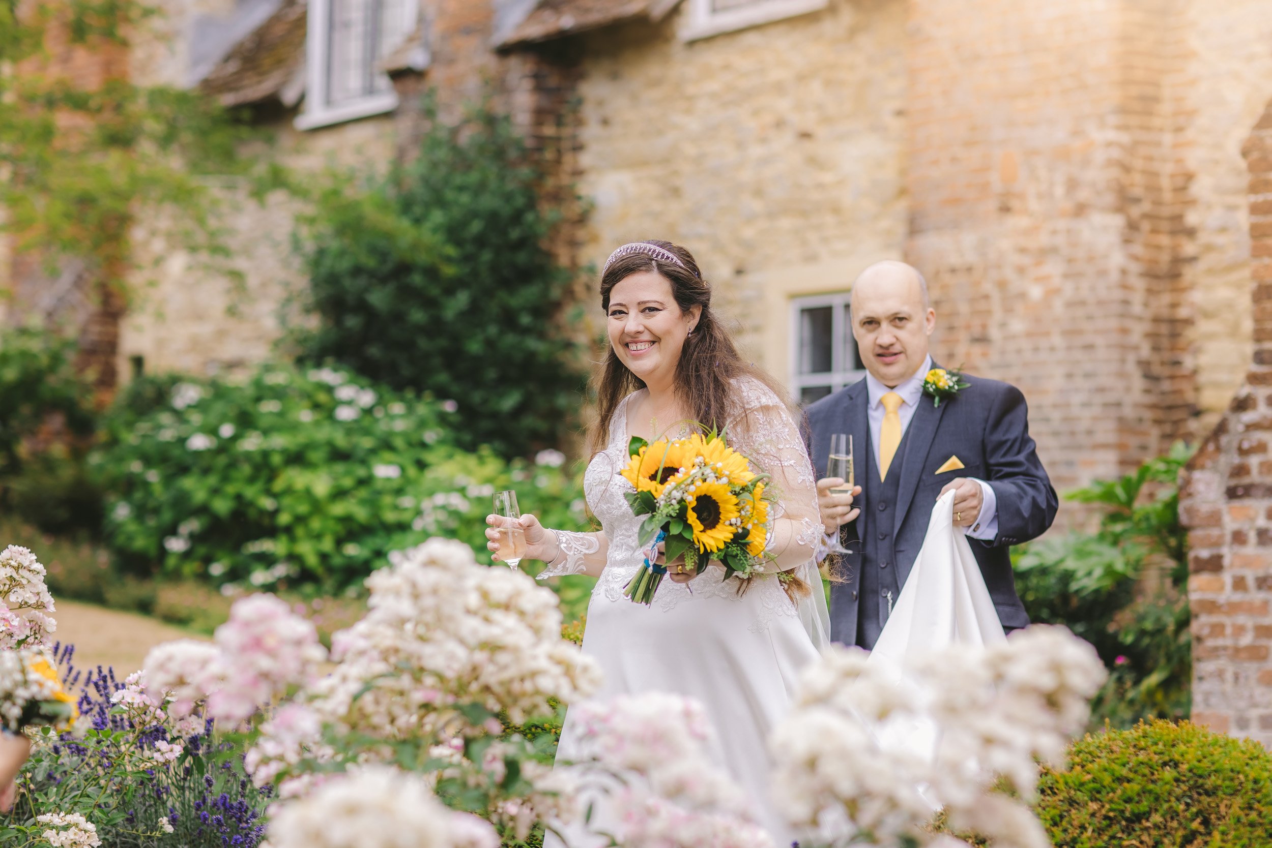 A smiling bride in a white wedding dress holding a bouquet of sunflowers, and a groom in a navy suit with a yellow tie, holding a glass of champagne and a white cloth, walking through a garden with pink and white flowers, in front of an old brick bui