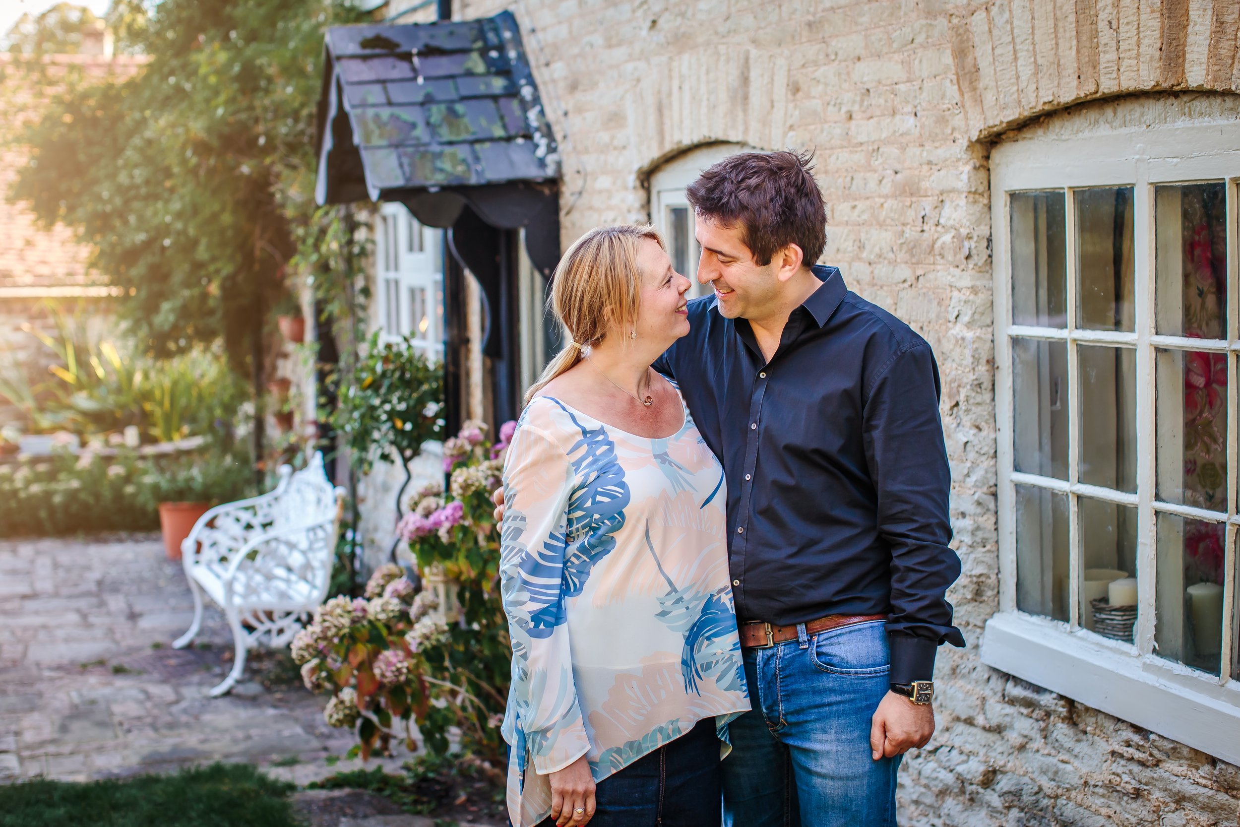 A couple smiling and looking at each other in front of a stone house with a window and a garden with flowers.