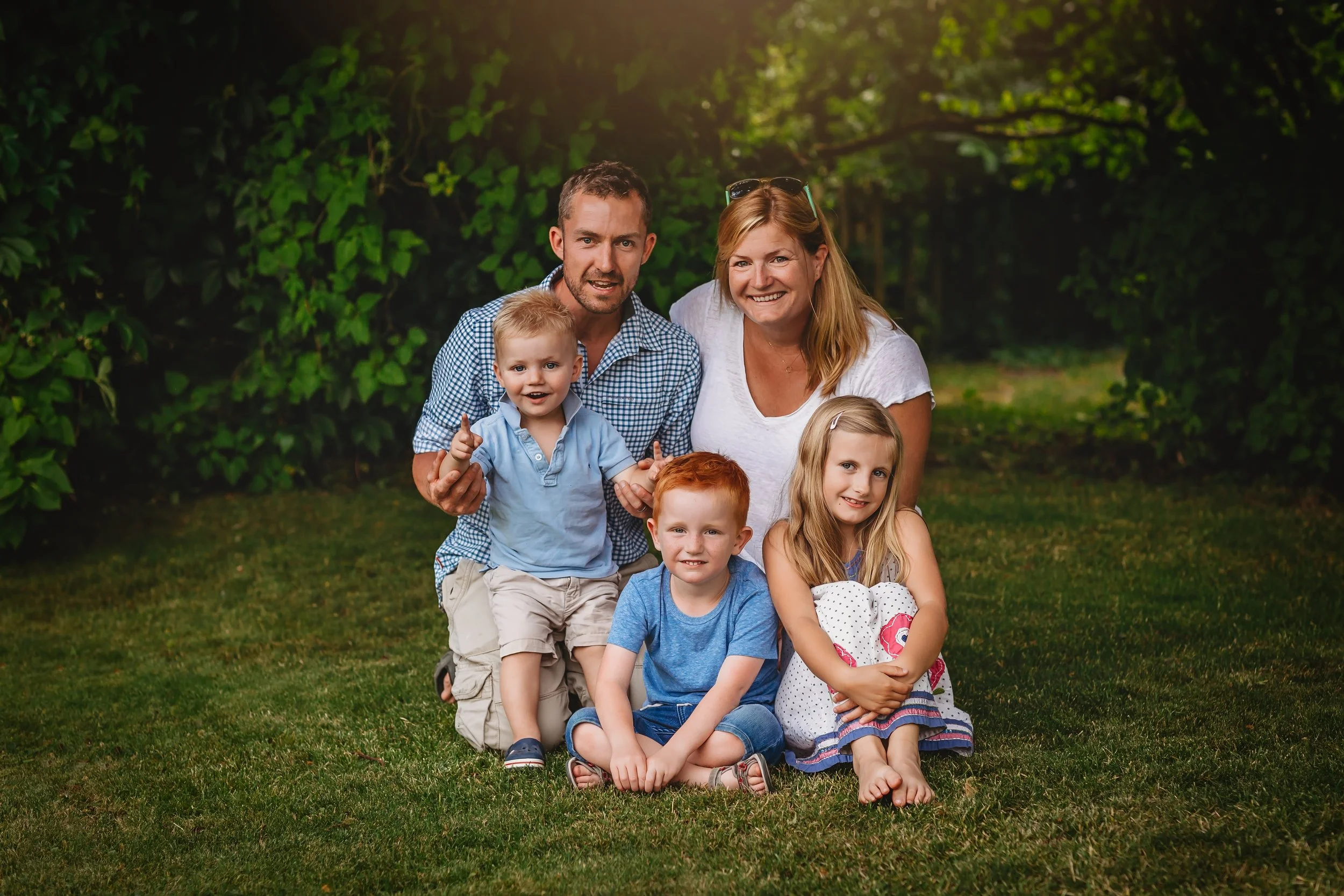 A happy family of six, including two parents and four children, sitting on grass in a park during daytime. They are smiling and looking at the camera, surrounded by green trees.