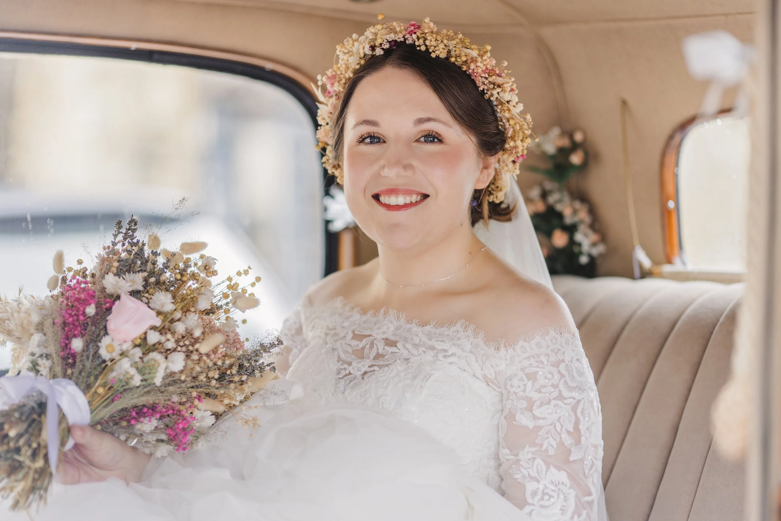 A bride with a floral headpiece sitting in a vintage vehicle, holding a bouquet of dried flowers, smiling at the camera.