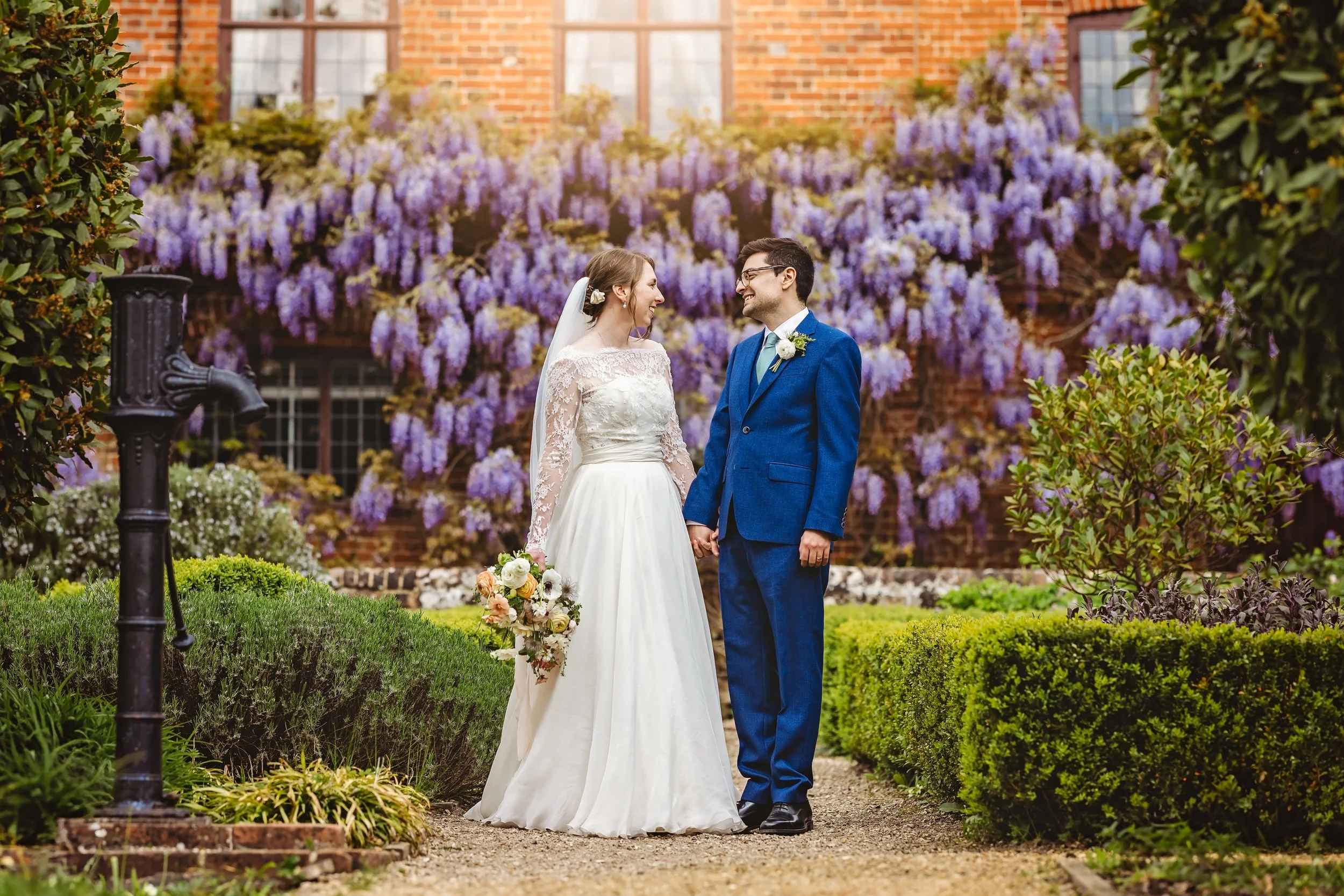 A bride and groom holding hands and smiling at each other in a garden with purple flowers and greenery.