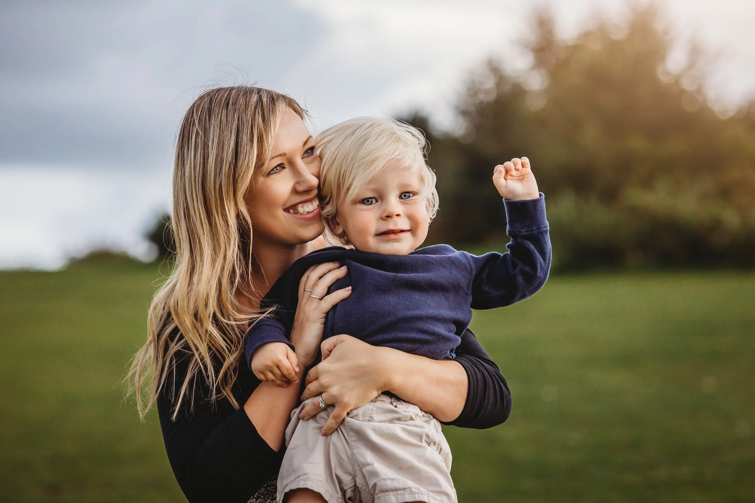 A woman with long blonde hair smiling while holding a young blonde-haired boy outdoors in a park with green grass and trees, as the boy raises his arm in a fist.