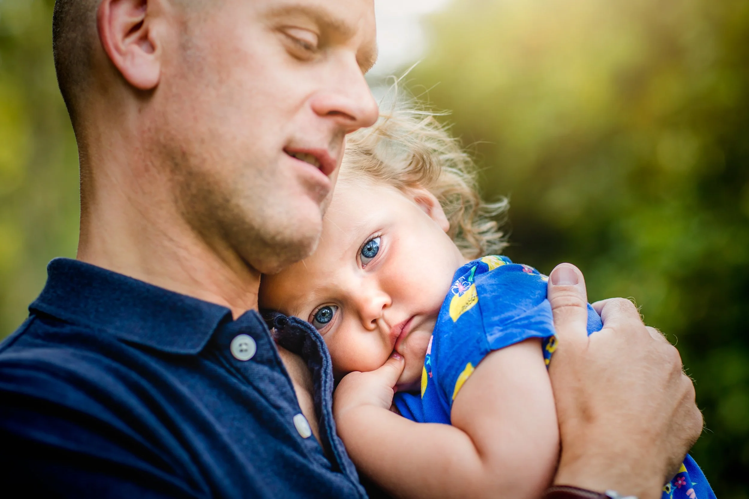 A man holding a young girl with blue eyes, wearing a blue shirt with colorful patterns, in an outdoor setting with blurred green background.