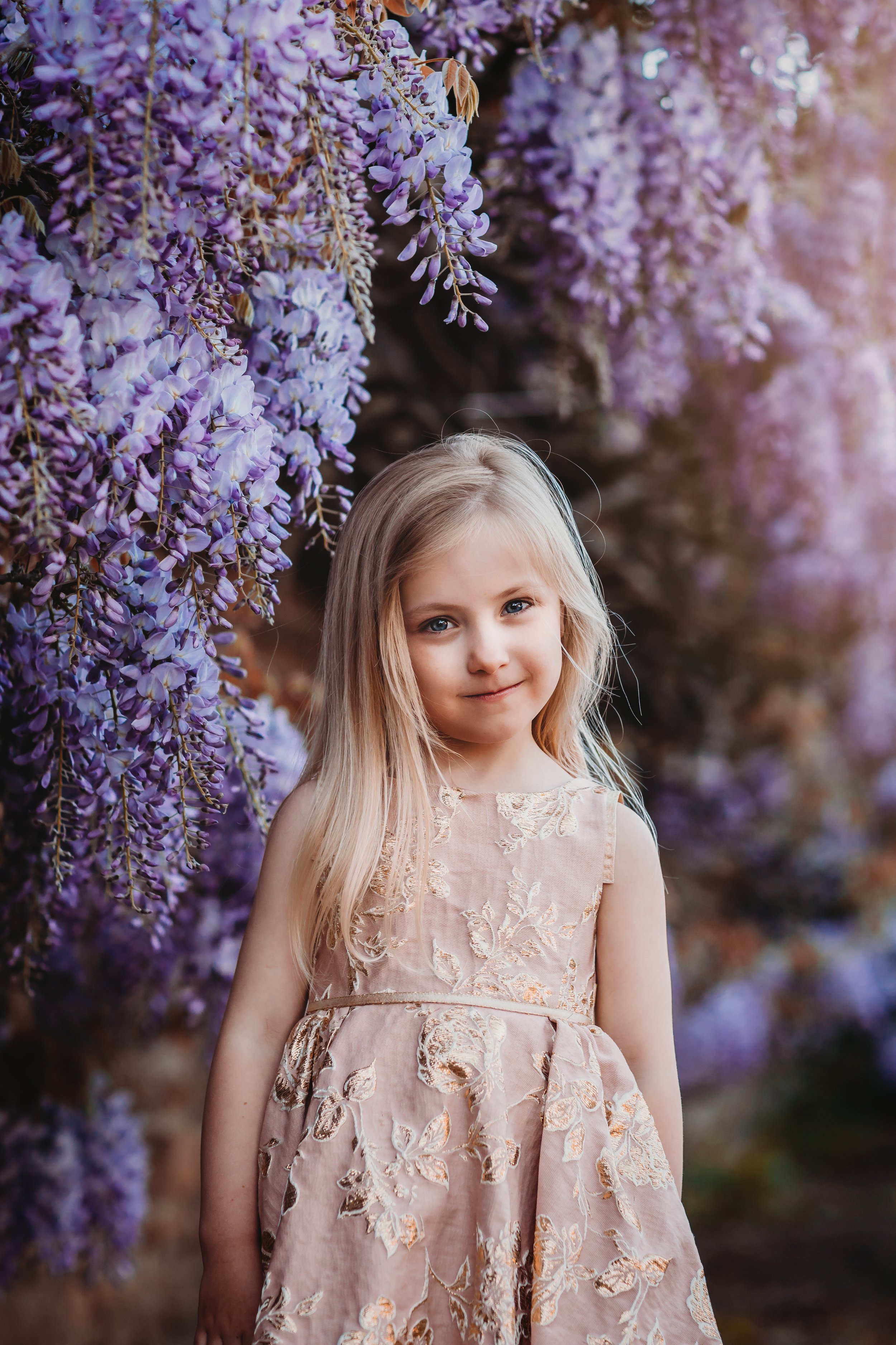 A young girl standing outdoors among purple wisteria flowers, wearing a light pink embroidered dress, with long blonde hair and a gentle smile.
