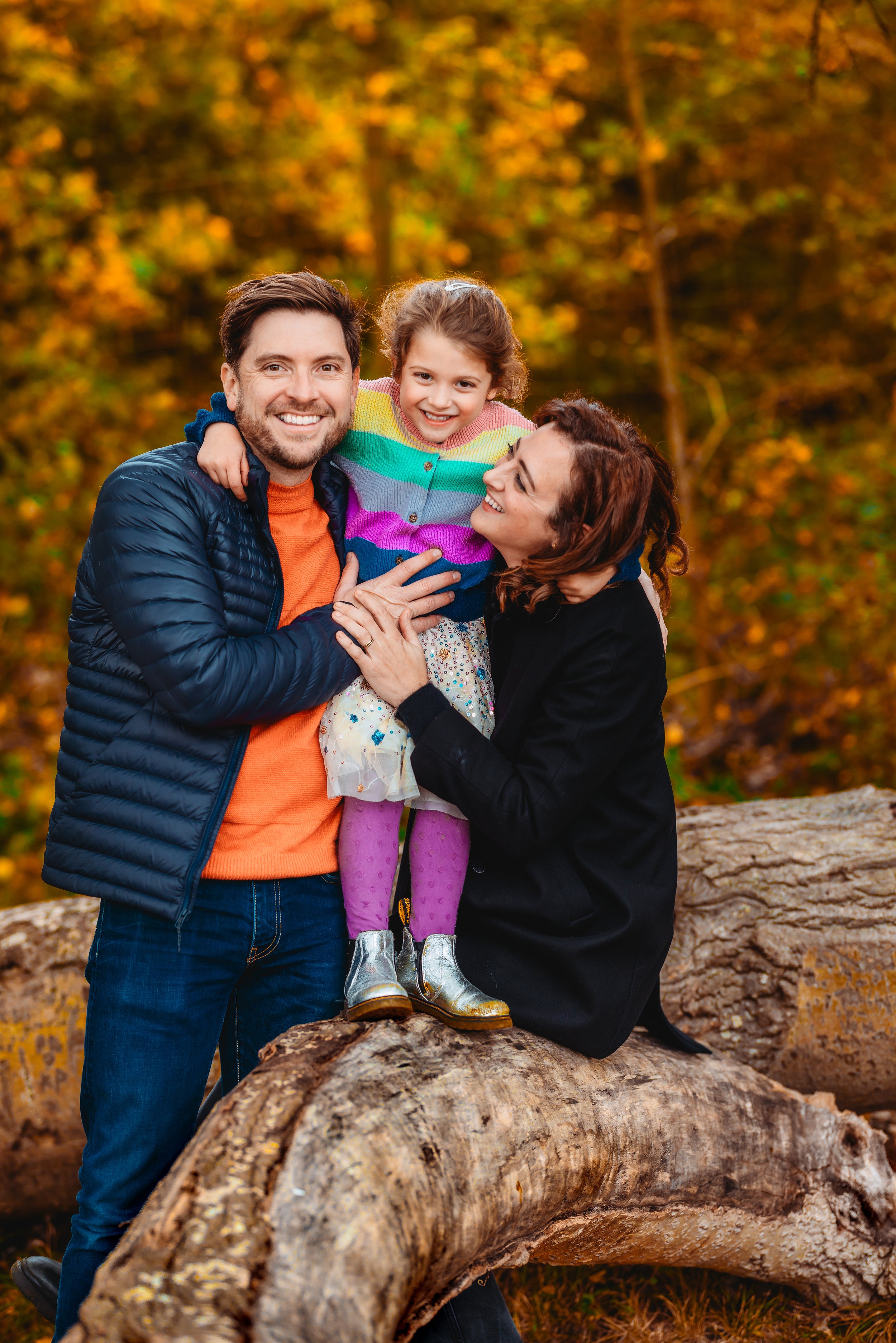 Family of three enjoying fall outdoors; father, mother, and young girl on a large fallen log, with colorful autumn leaves in background.