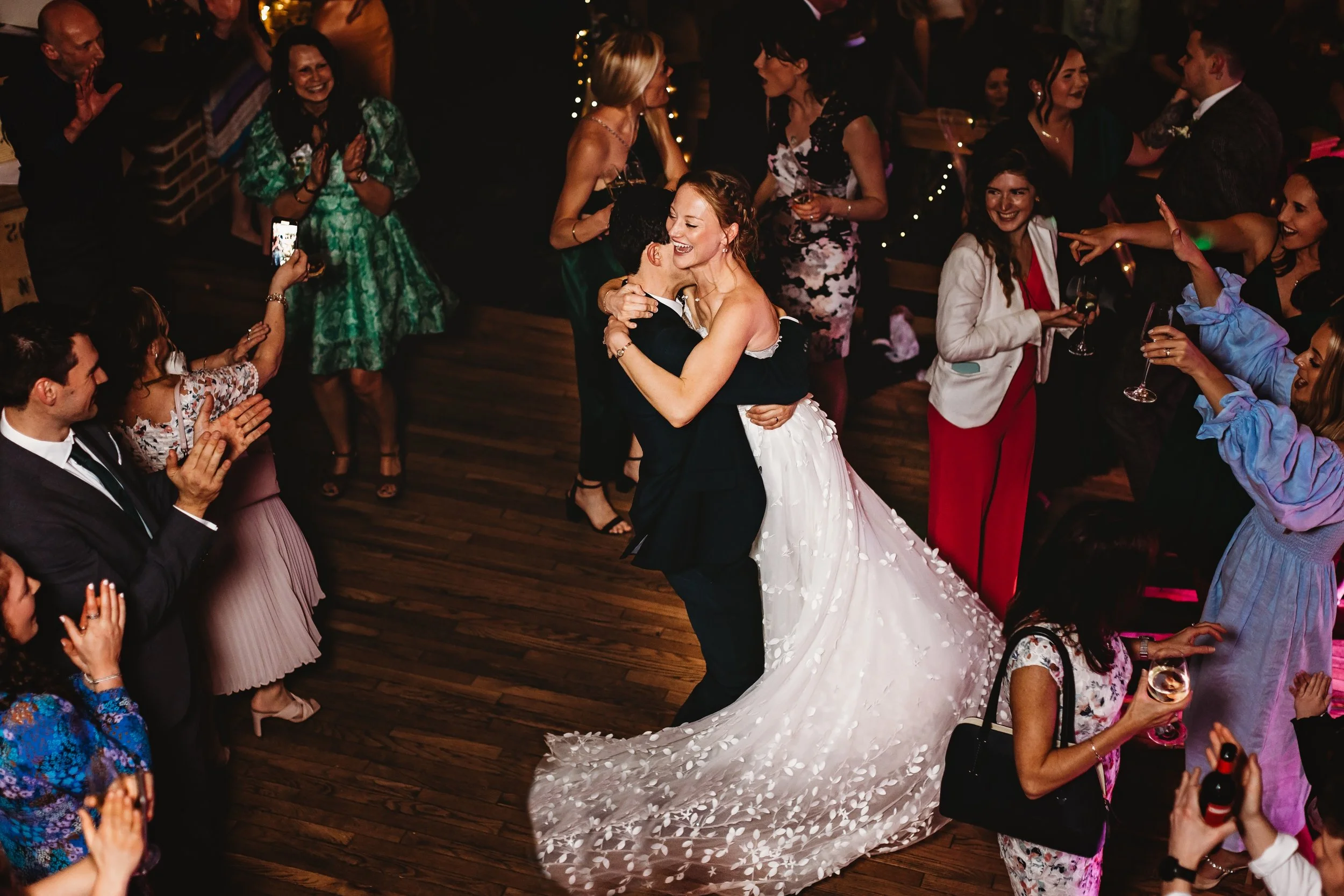 A bride and groom embrace during their wedding reception while guests surround them on the dance floor, clapping, smiling, and taking photos.