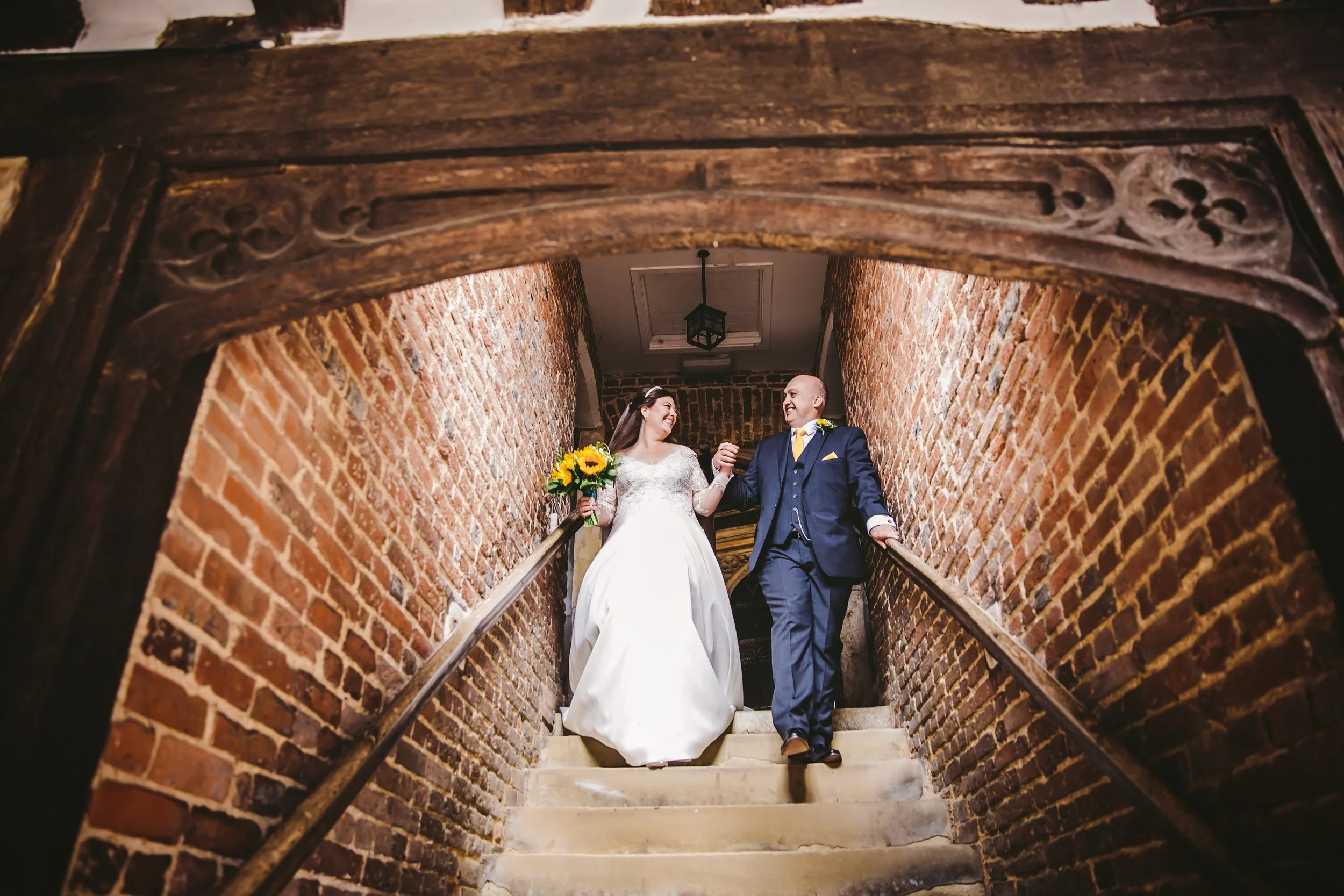 A bride and groom are walking down a staircase holding hands, smiling at each other. The bride is wearing a white wedding dress and holding a bouquet of sunflowers, while the groom is in a dark suit with a yellow tie. They are enclosed by brick walls