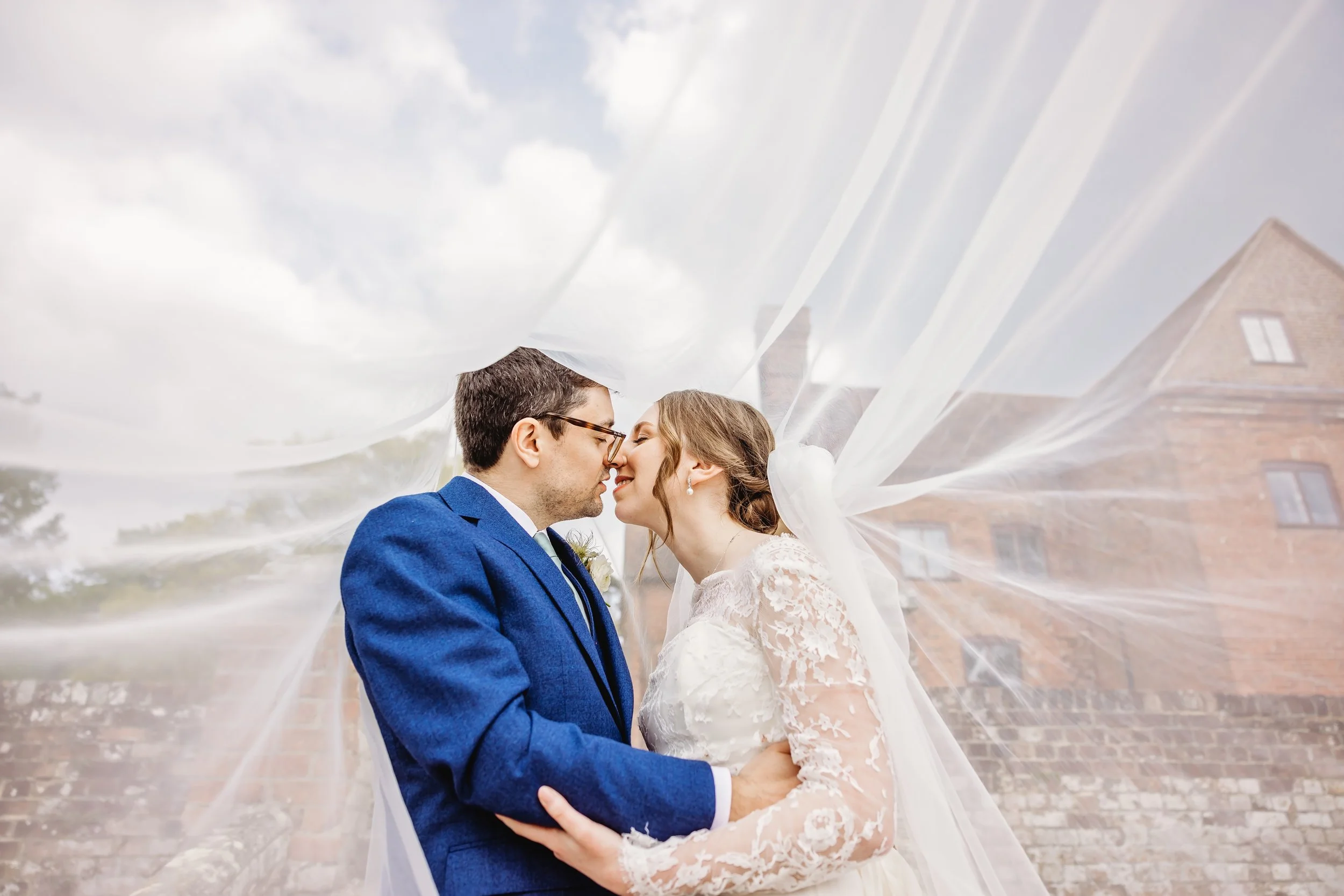 A bride and groom stand close together, about to kiss, outdoors on a cloudy day, with the bride's veil flowing around them and brick buildings in the background.