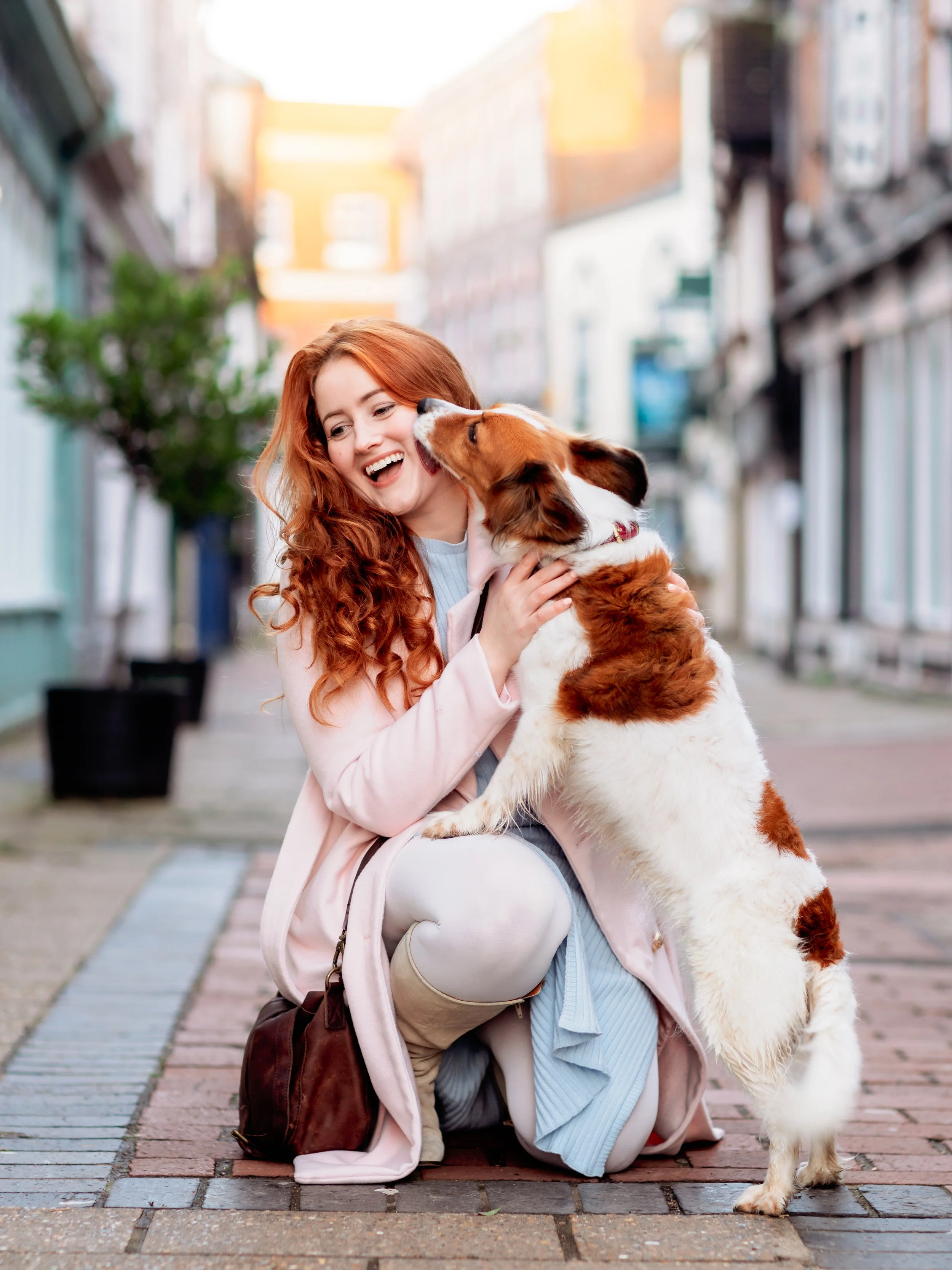 A young woman with long curly red hair kneels on a city sidewalk, smiling as a large brown and white dog licks her face. The woman is wearing a light pink coat, a light blue dress, and knee-high boots, and she has a brown handbag on the ground next t