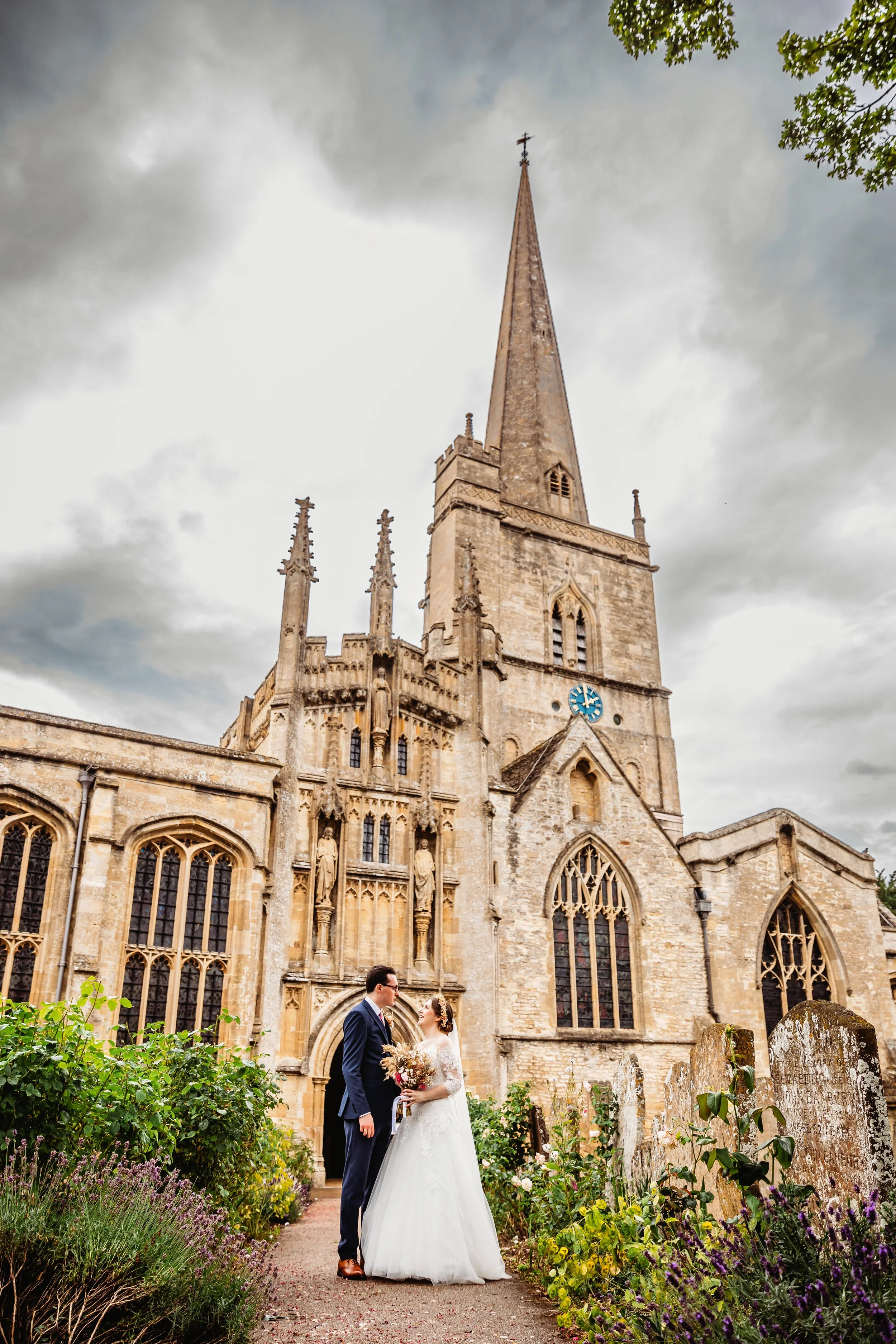 A bride and groom in wedding attire are standing in front of a large gothic-style church, holding a bouquet of flowers, surrounded by greenery and gravestones.
