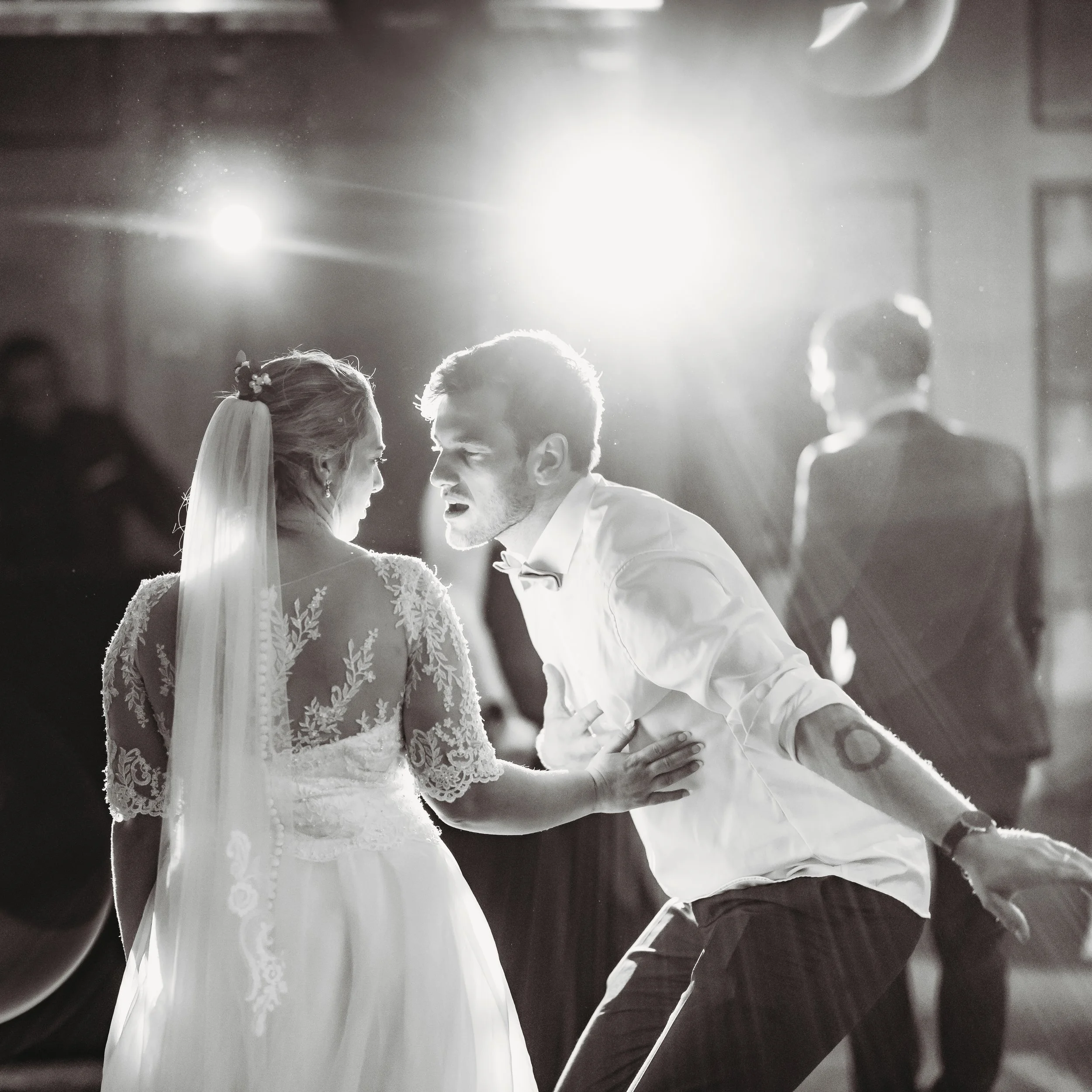 A black and white photograph of a wedding reception dance. A bride in a lace wedding gown and veil is dancing closely with a groom in a white shirt, who is leaning forward and singing or speaking. There are bright stage lights behind them and other g