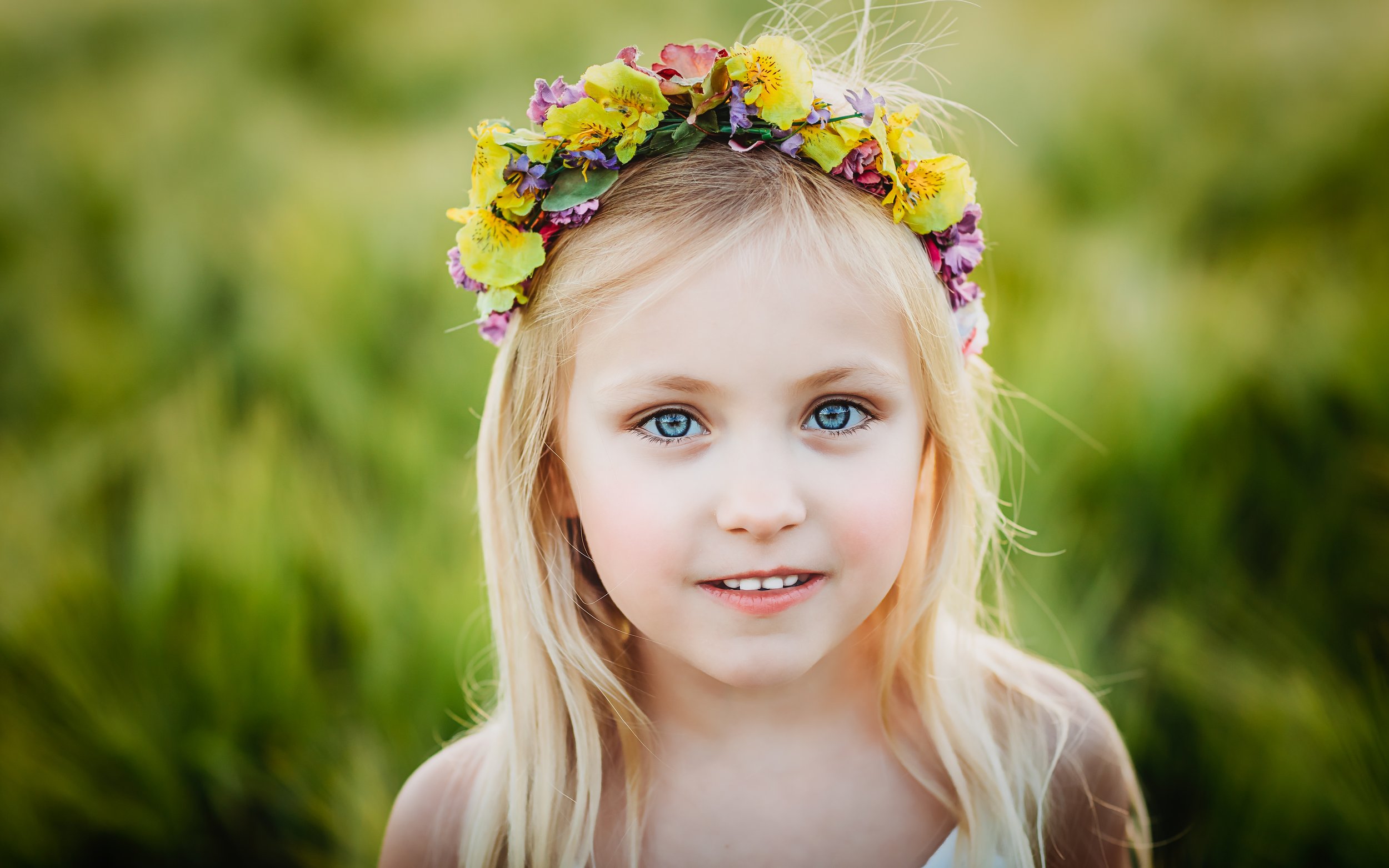 A young girl with long blonde hair and blue eyes wearing a colorful flower crown outdoors.